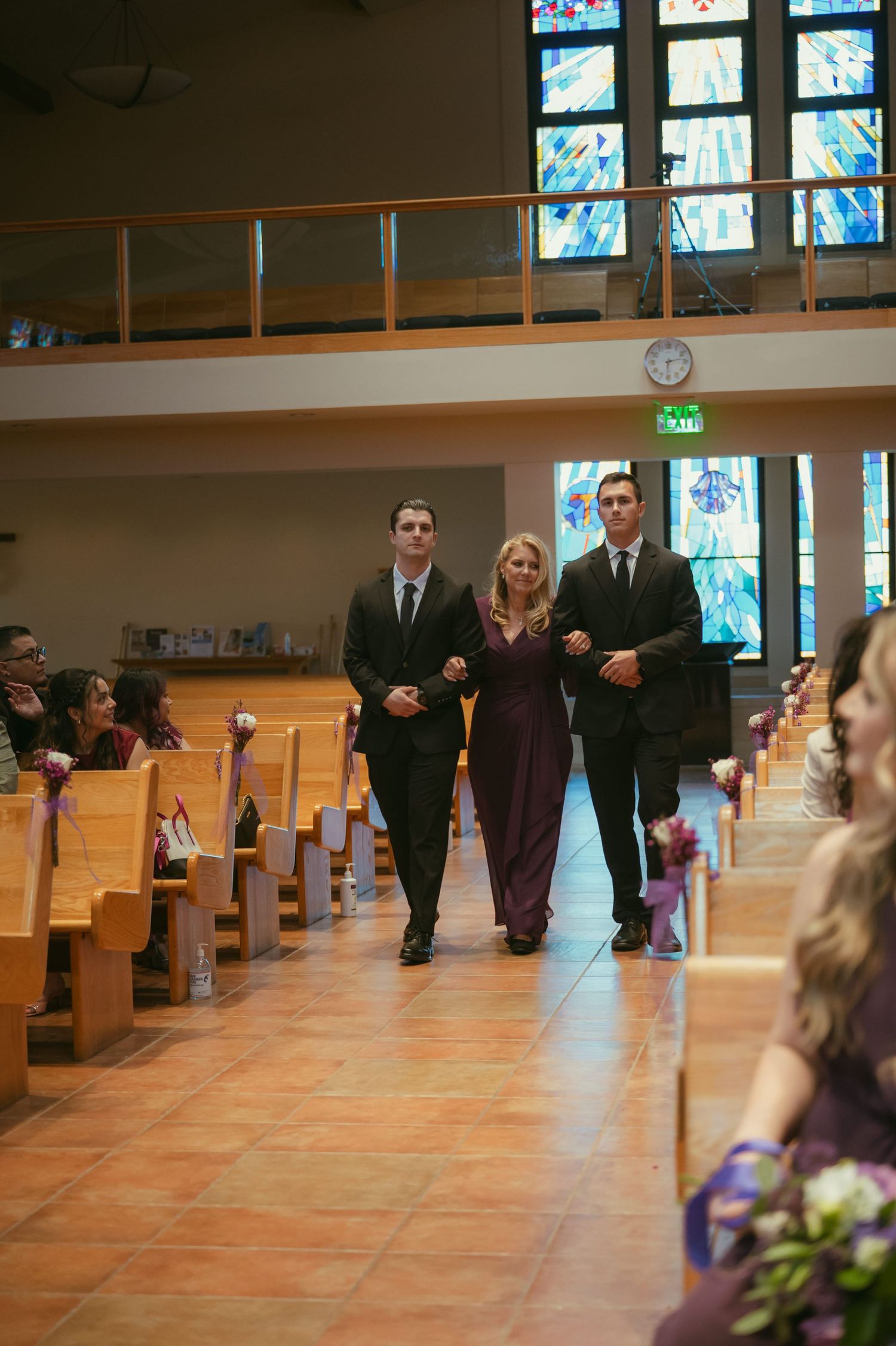 Woman escorted by two men in black suits down church aisle during wedding ceremony with stained glass windows.