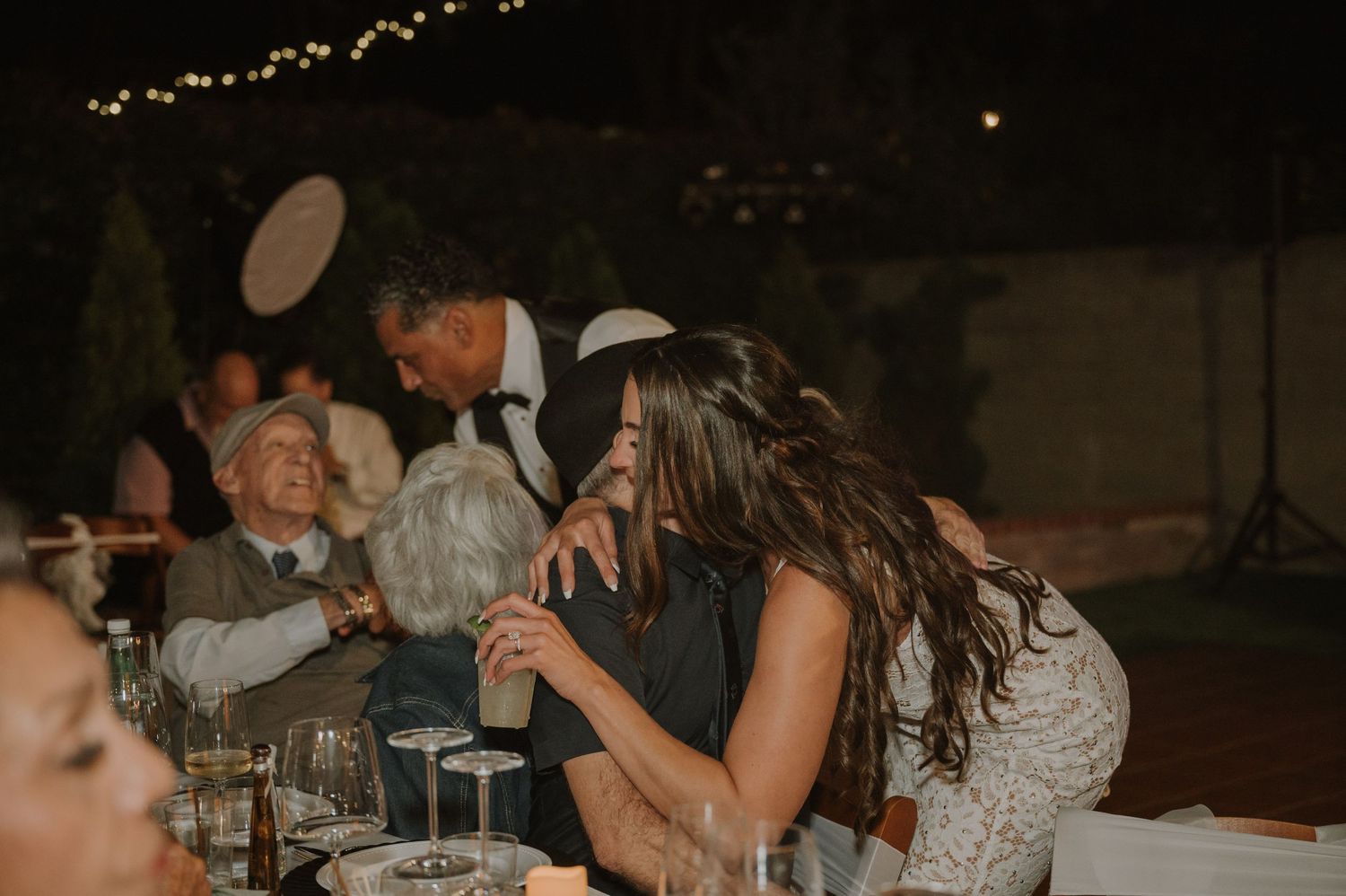 Guests laughing and celebrating at an outdoor evening wedding reception dinner table.