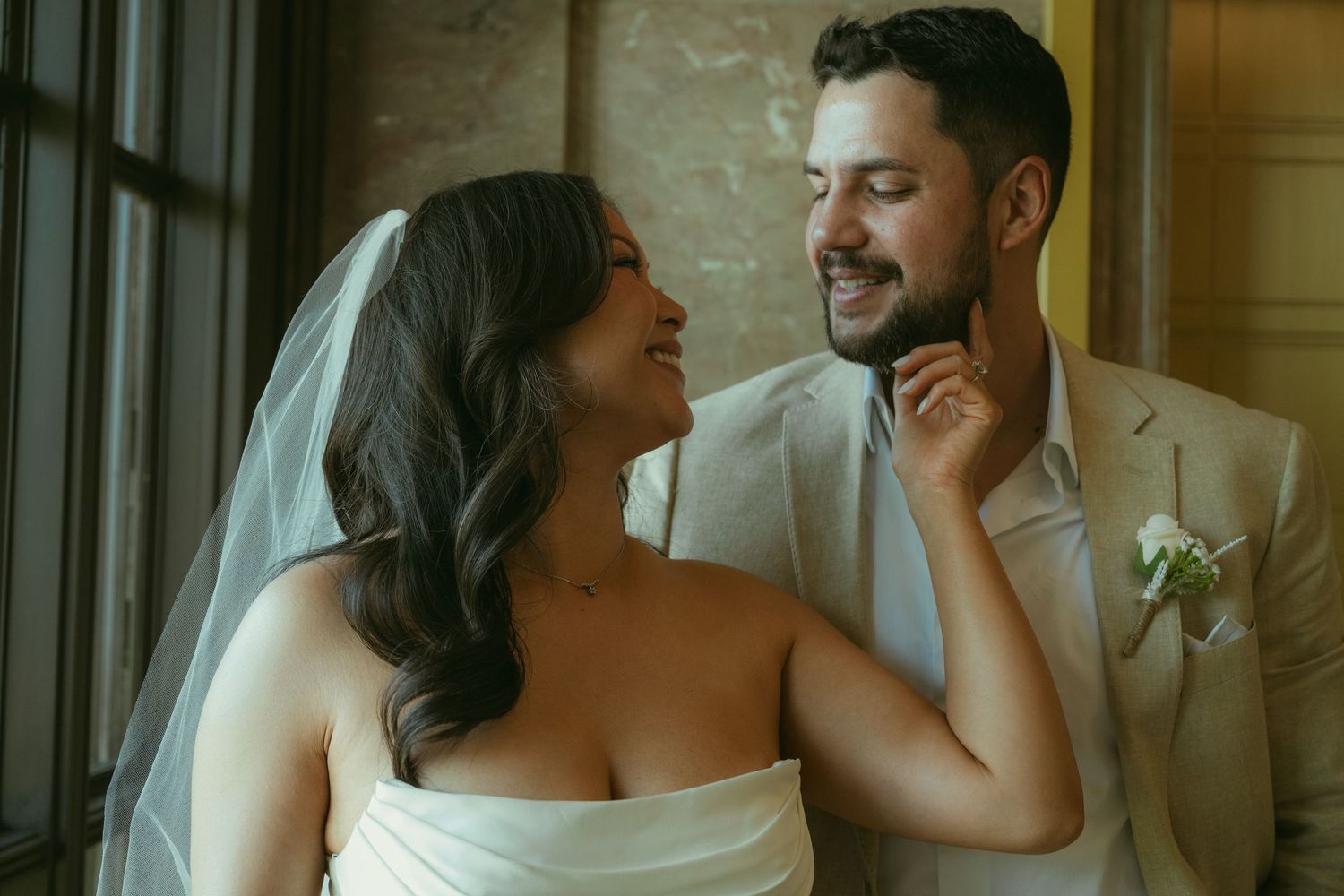 A smiling bride in a white strapless gown and veil gazes at her groom in a tan suit at their wedding.