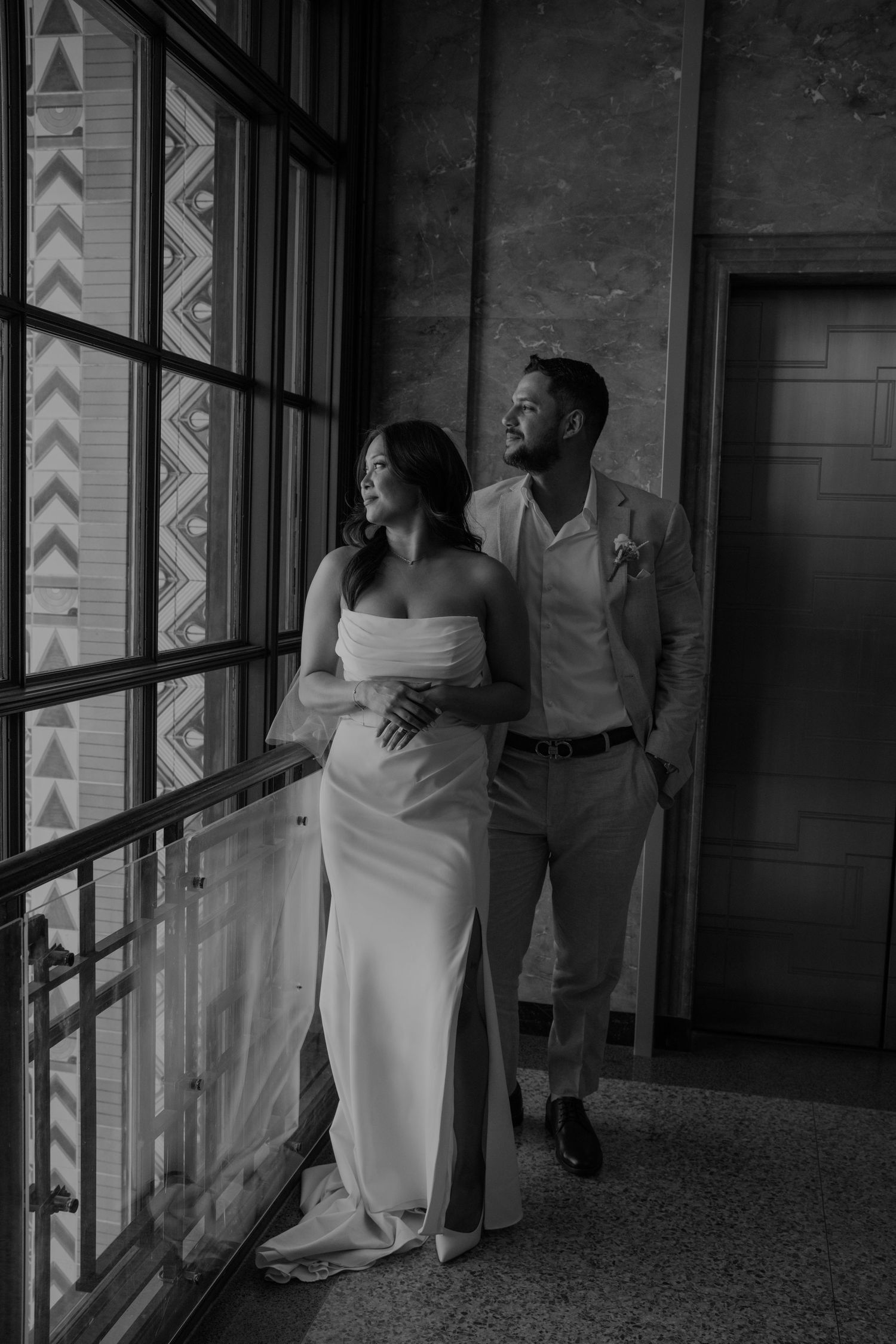 Black and white photo of a bride and groom standing by a large window, gazing out together on their wedding day.