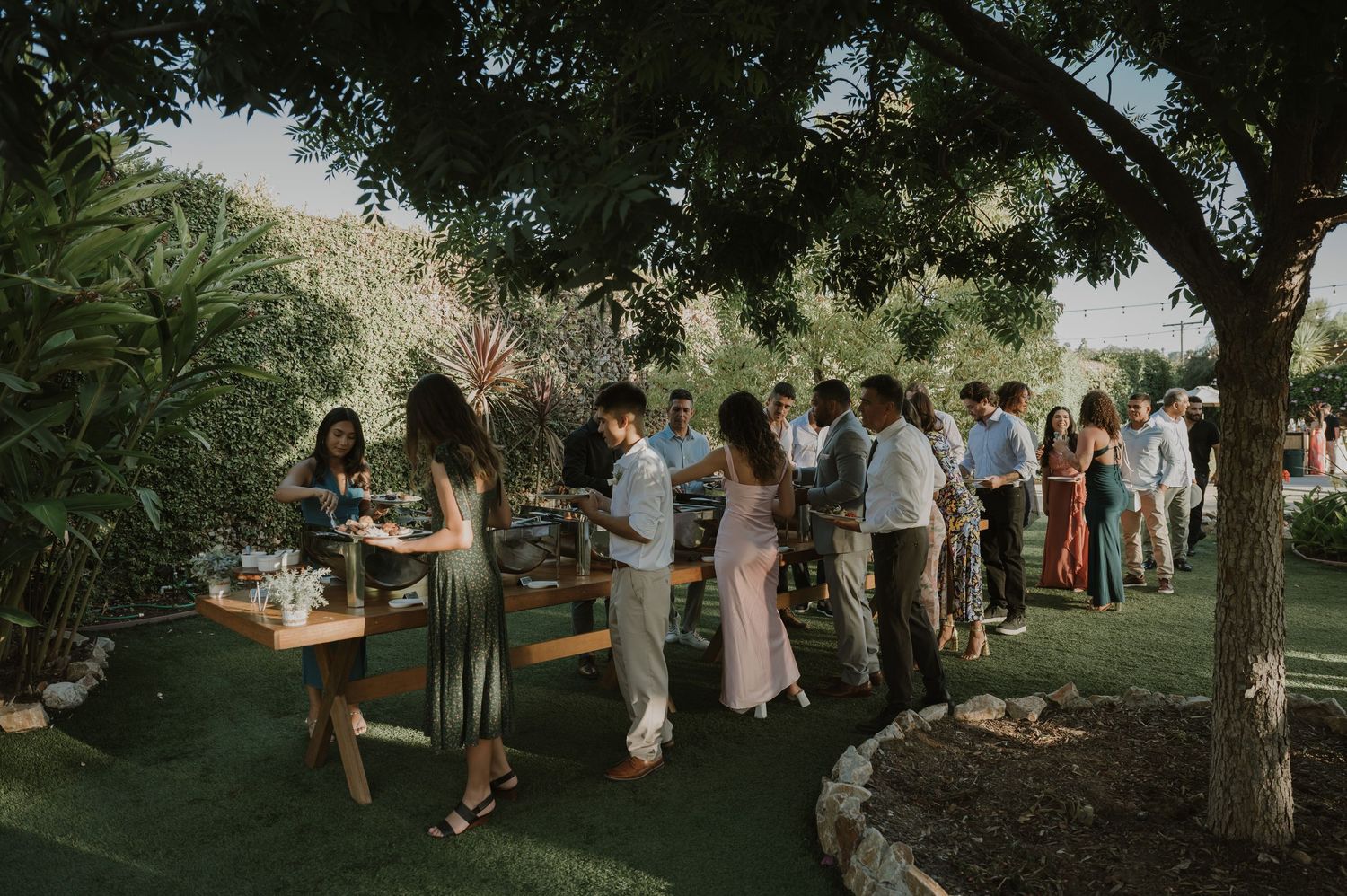 Wedding guests mingle and line up at an outdoor reception surrounded by lush garden greenery.