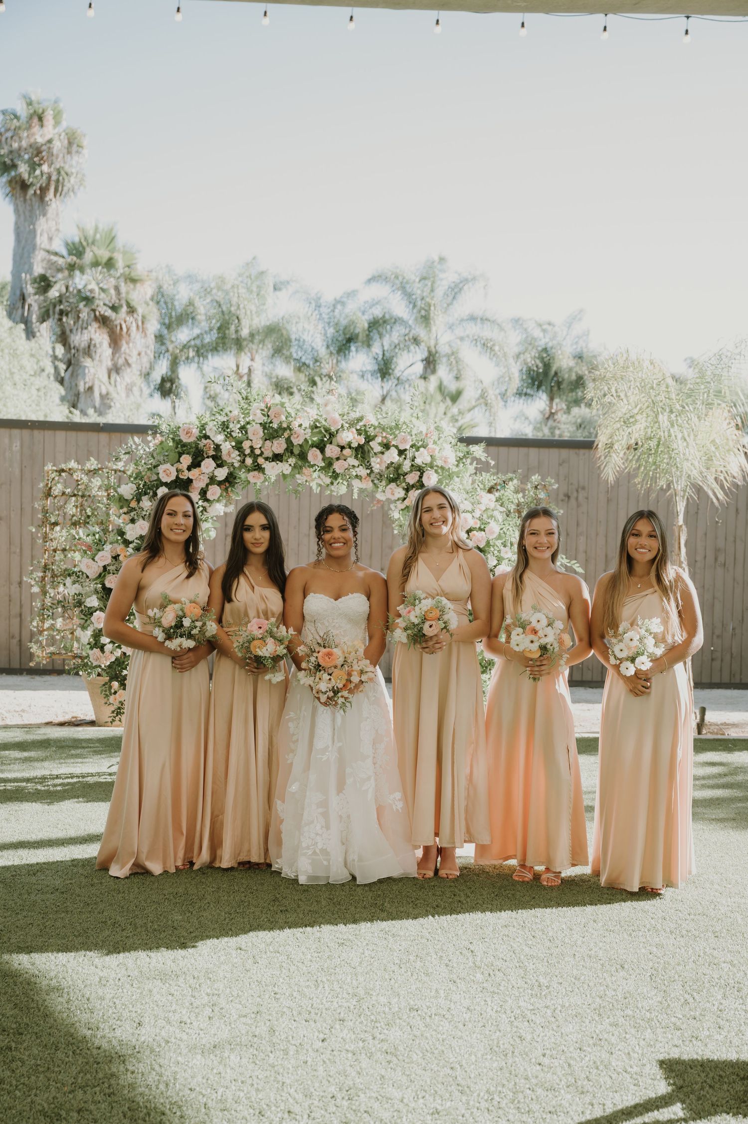 Smiling bride with bouquet stands with five bridesmaids in blush gowns before romantic floral backdrop.