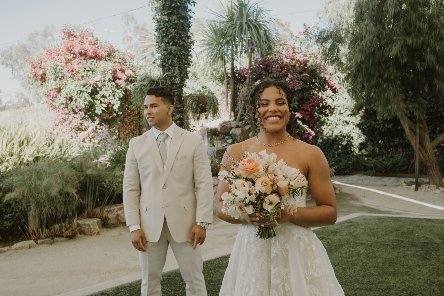 Bride in lace gown holds bouquet while groom stands behind her in a garden.