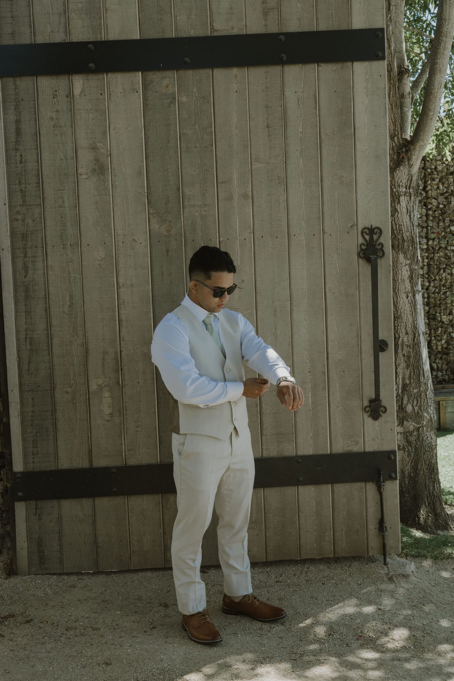 Groom in white suit and sunglasses adjusting cufflinks beside rustic wooden barn doors on wedding day.