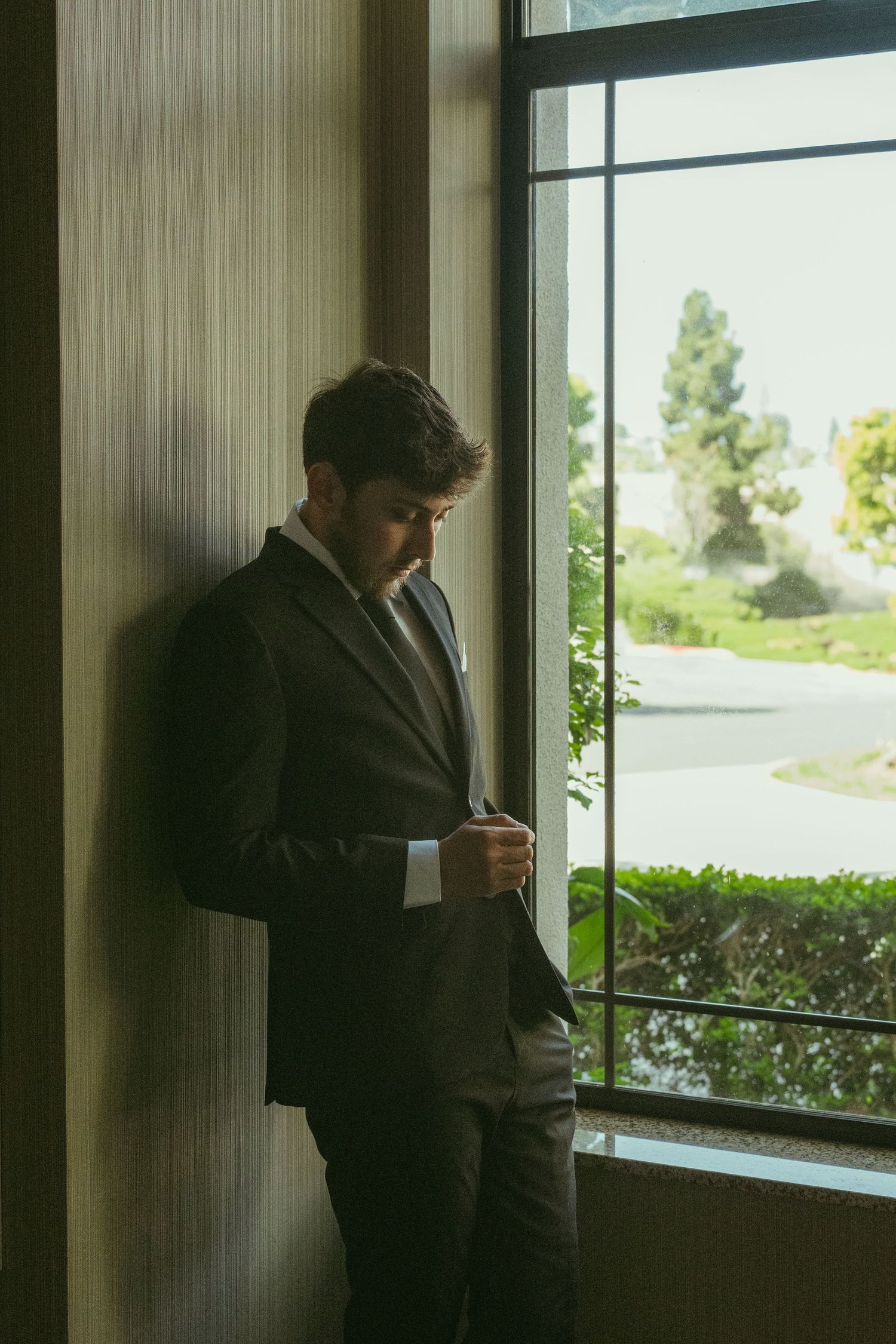 A well-dressed man in a dark suit adjusts his jacket while standing by a large window with a green outdoor view.