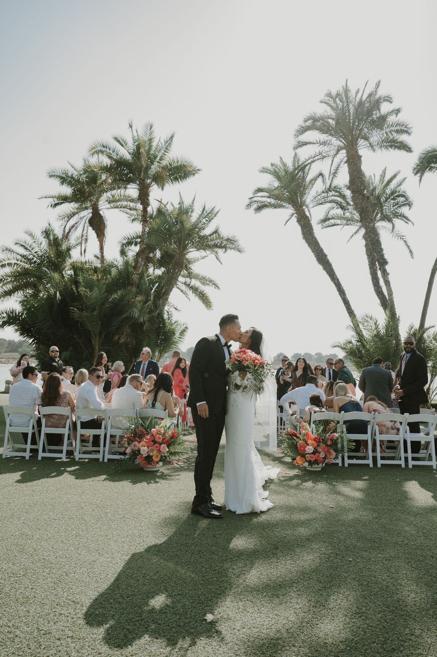Outdoor wedding ceremony under palm trees with white chairs and coral flower arrangements.
