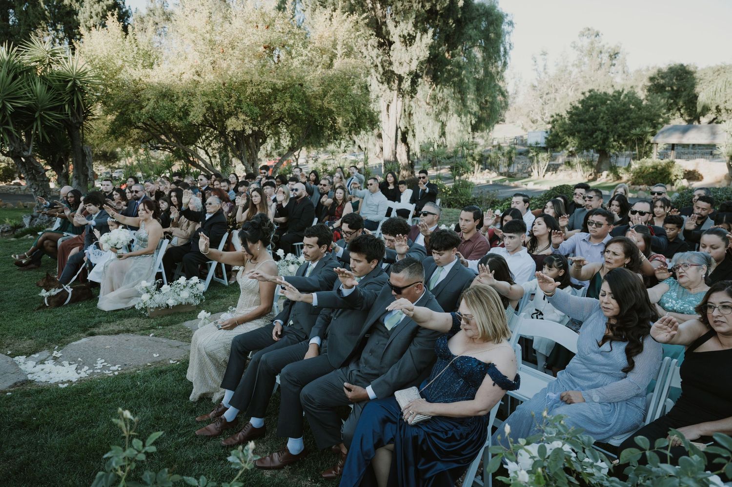 Panoramic view of wedding guests seated outdoors under trees during a daytime ceremony.