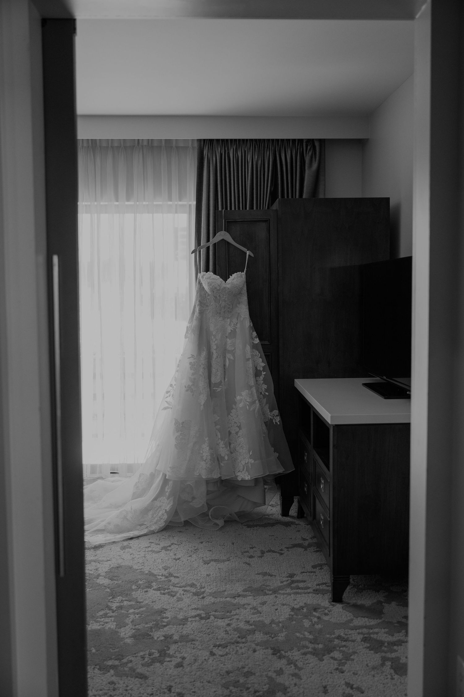Elegant white lace wedding dress hanging in a room with sheer curtains and dark furniture.