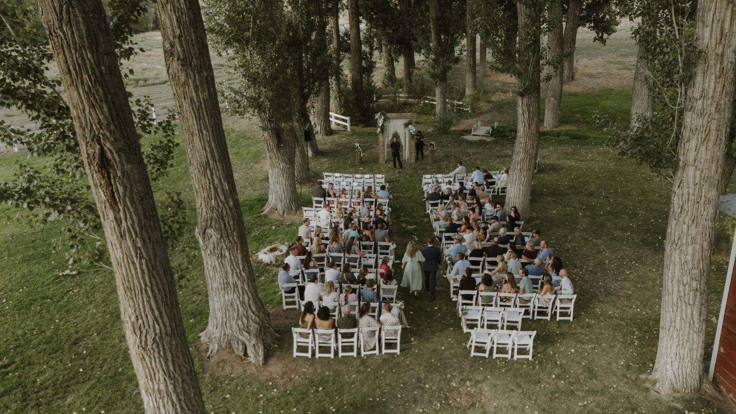 Panoramic view of an outdoor wedding ceremony space with white chairs arranged between tall trees.