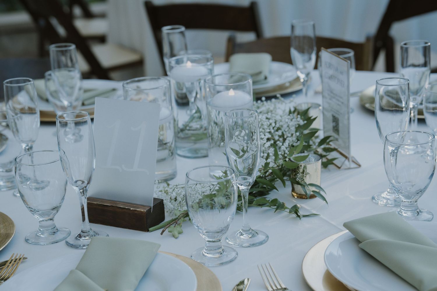 Wedding reception tables decorated with white linens, glassware, and delicate greenery centerpieces.