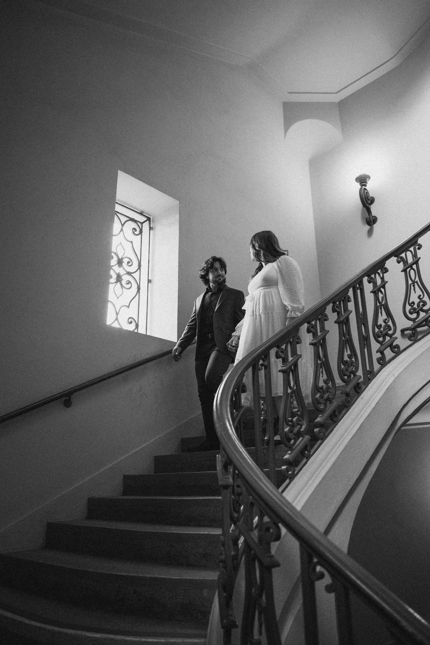 A couple ascending an elegant staircase with ornate iron railings in black and white.