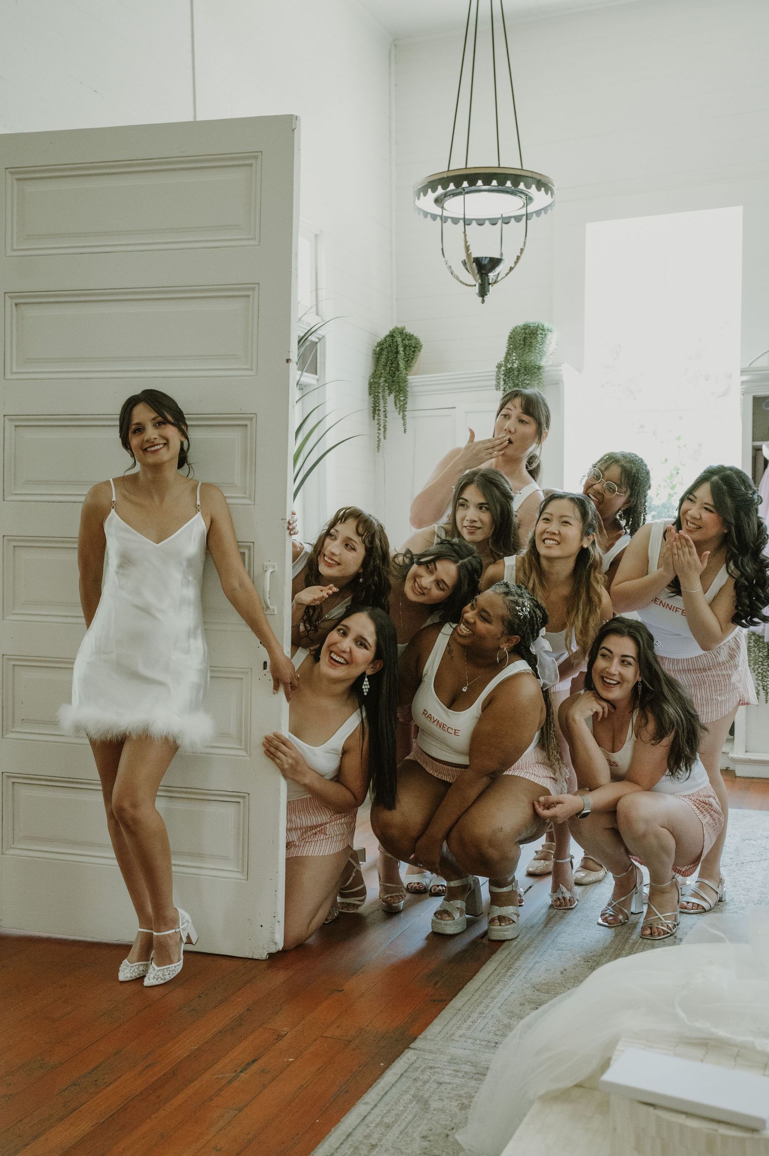 Bride in white mini dress poses with smiling bridesmaids in pink and white outfits in elegant room.