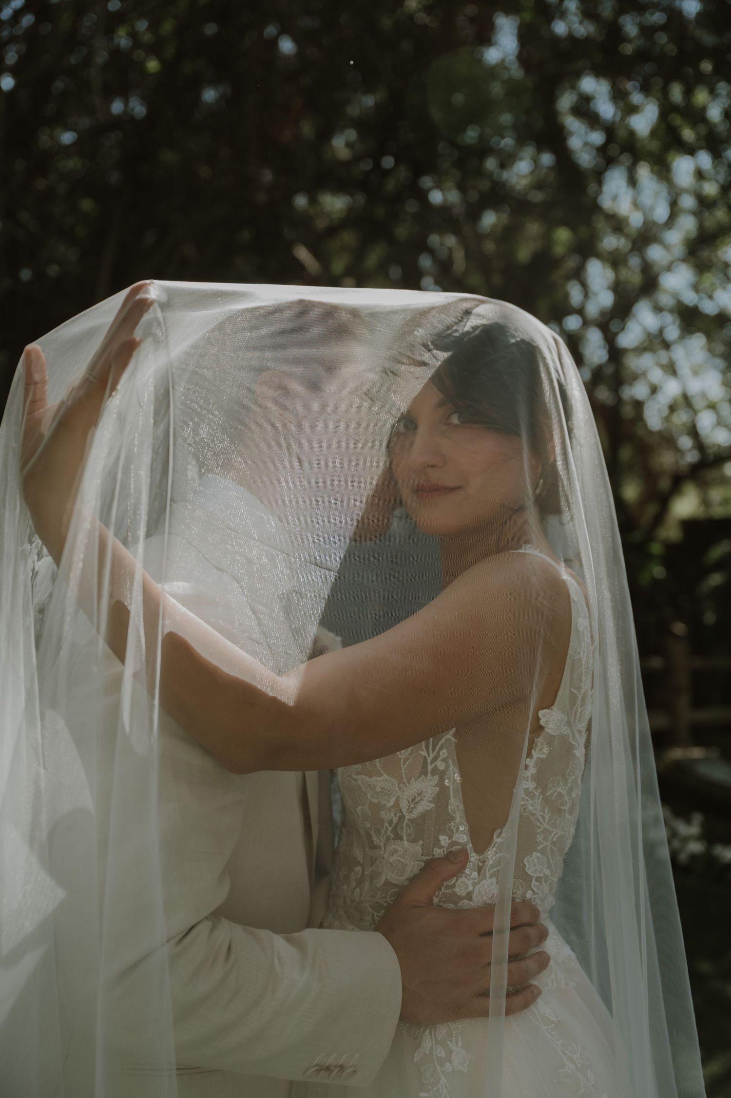 Bride and groom share a romantic embrace under a flowing white wedding veil outdoors.