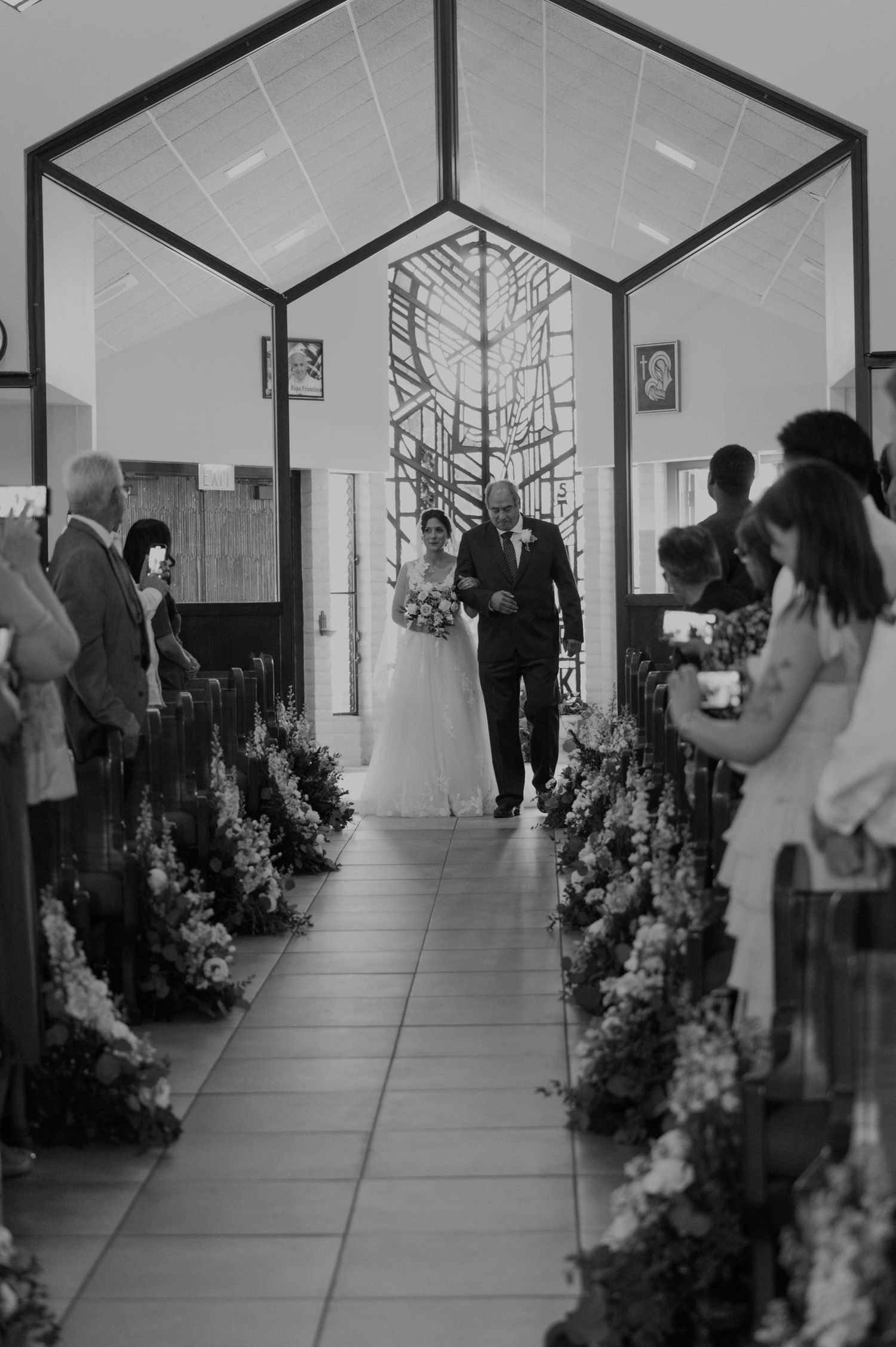 Black and white photo of bride walking down church aisle with escort, flanked by floral arrangements and wedding guests.