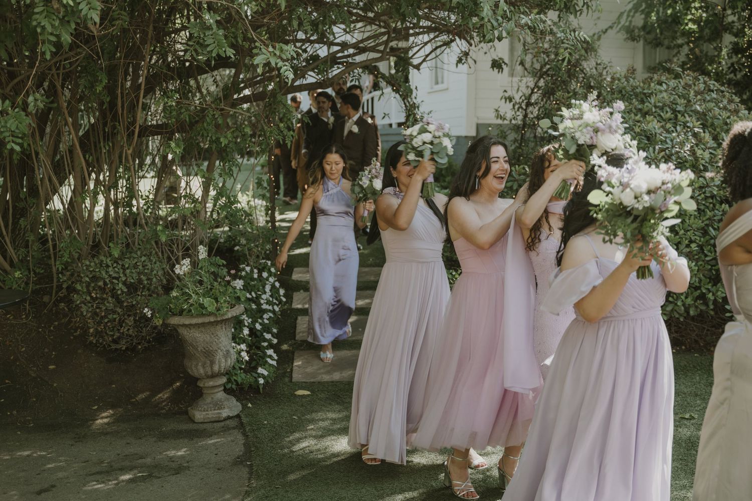 Bridesmaids in soft pink and lavender gowns walk through a lush garden carrying white floral bouquets at a wedding.