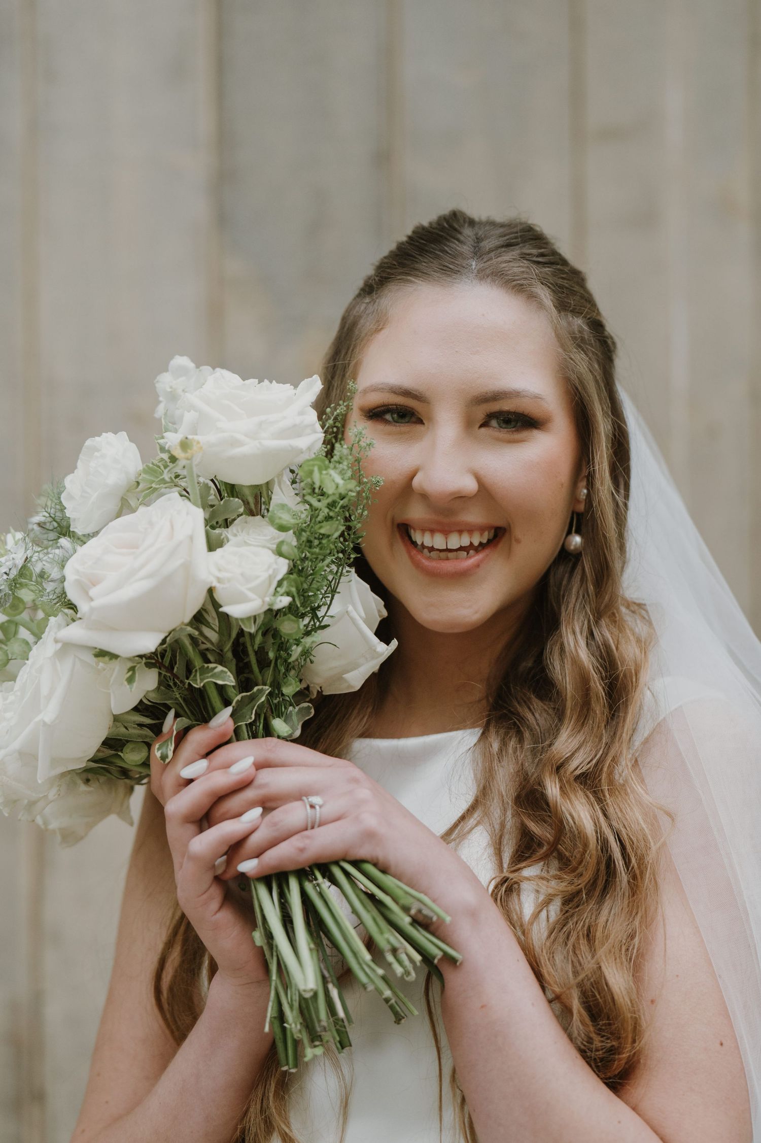 Radiant bride smiles holding white rose bouquet with greenery, wearing veil and wedding ring.