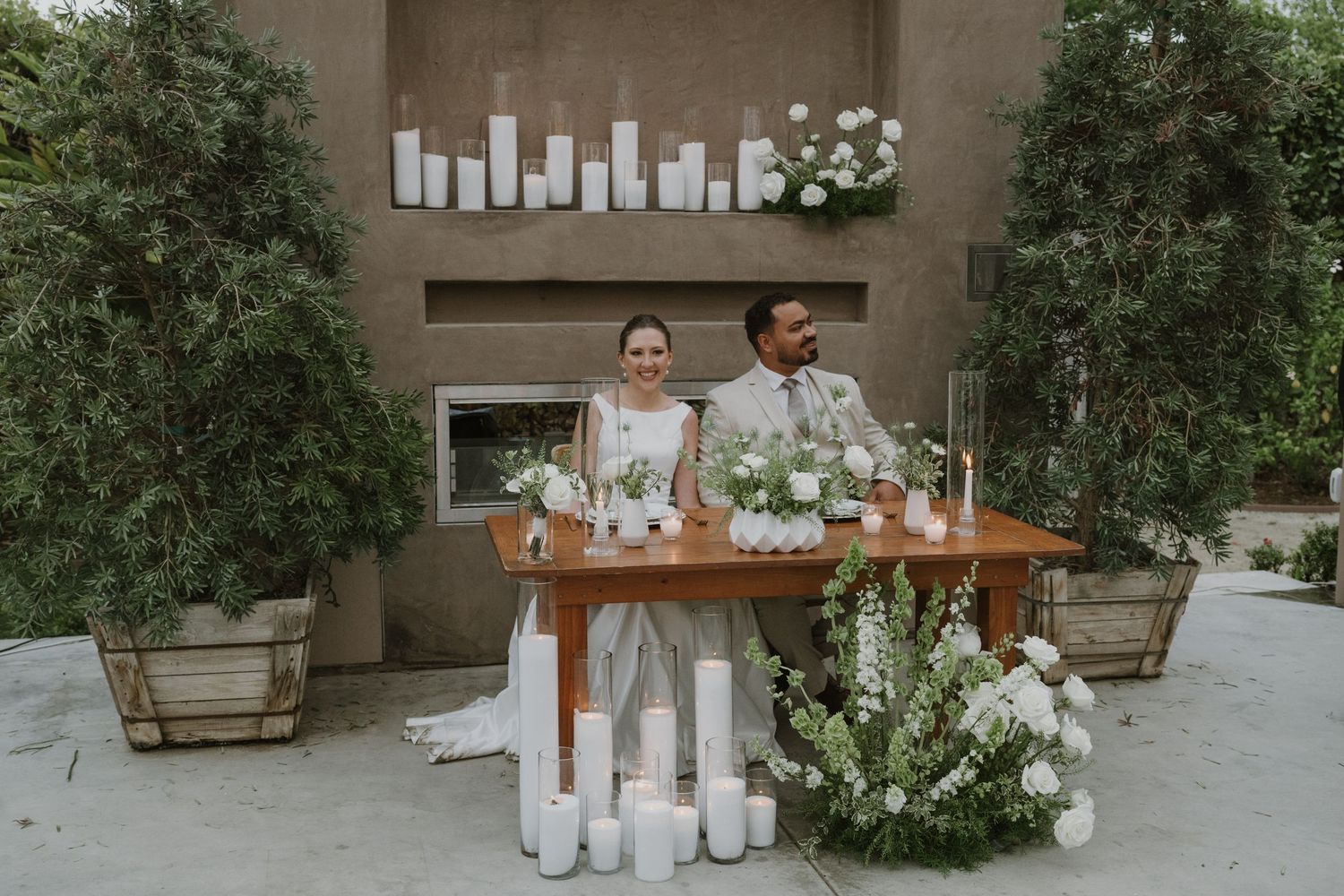 Bride and groom seated at floral wedding sweetheart table with white candles and greenery backdrop outdoors.