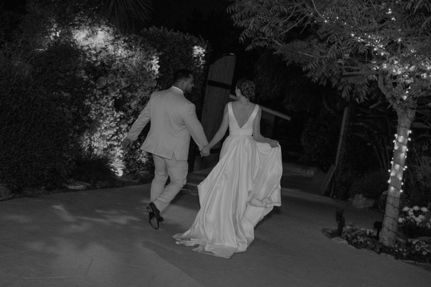 Black and white photo of bride and groom holding hands walking through garden with twinkling fairy lights.