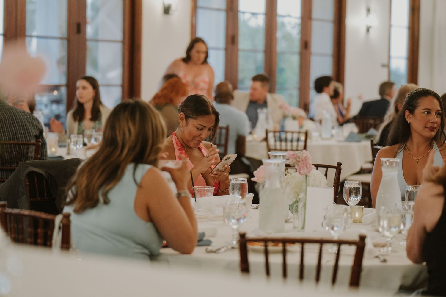 Wedding reception guests seated at elegantly decorated tables with floral centerpieces and glassware.
