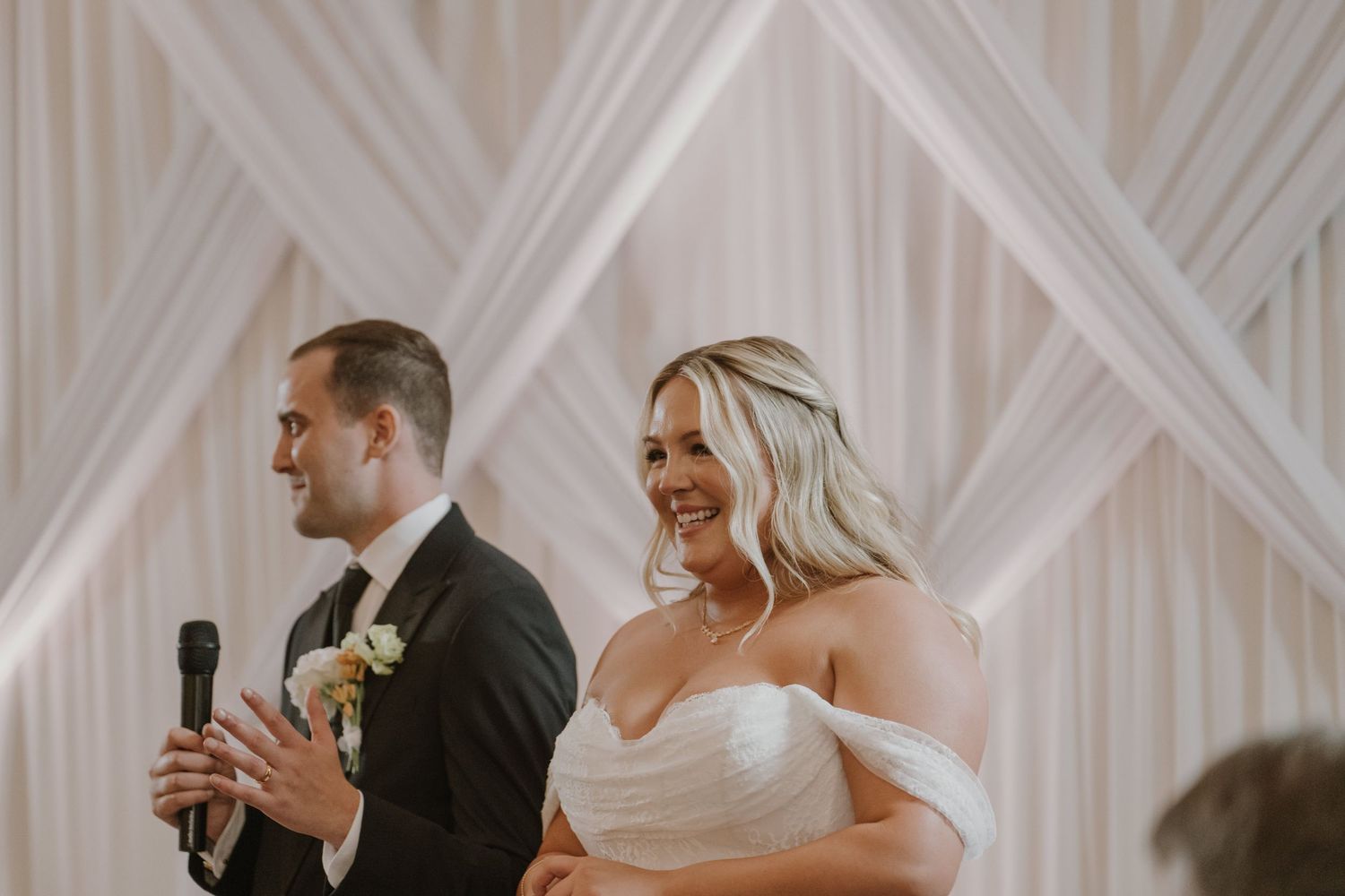 Bride and groom smiling and laughing together during wedding reception speech with elegant white drape backdrop.