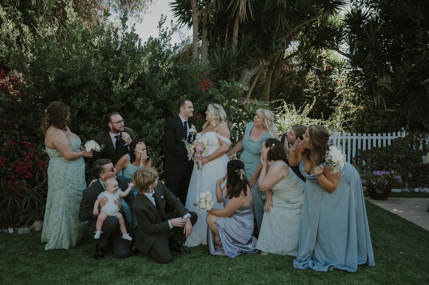 Wedding party group photo with bride, groom, bridesmaids in blue dresses gathered on garden lawn.