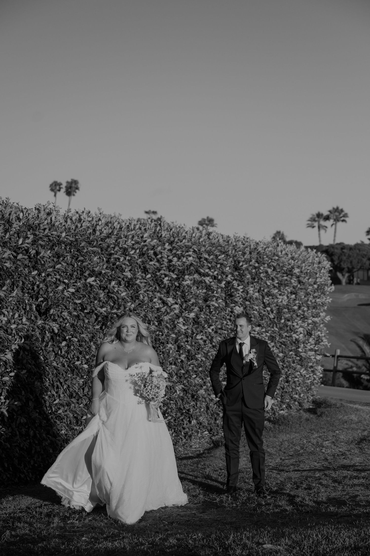 Bride and groom walking outdoors near a hedge with palm trees in background, black and white wedding photo.
