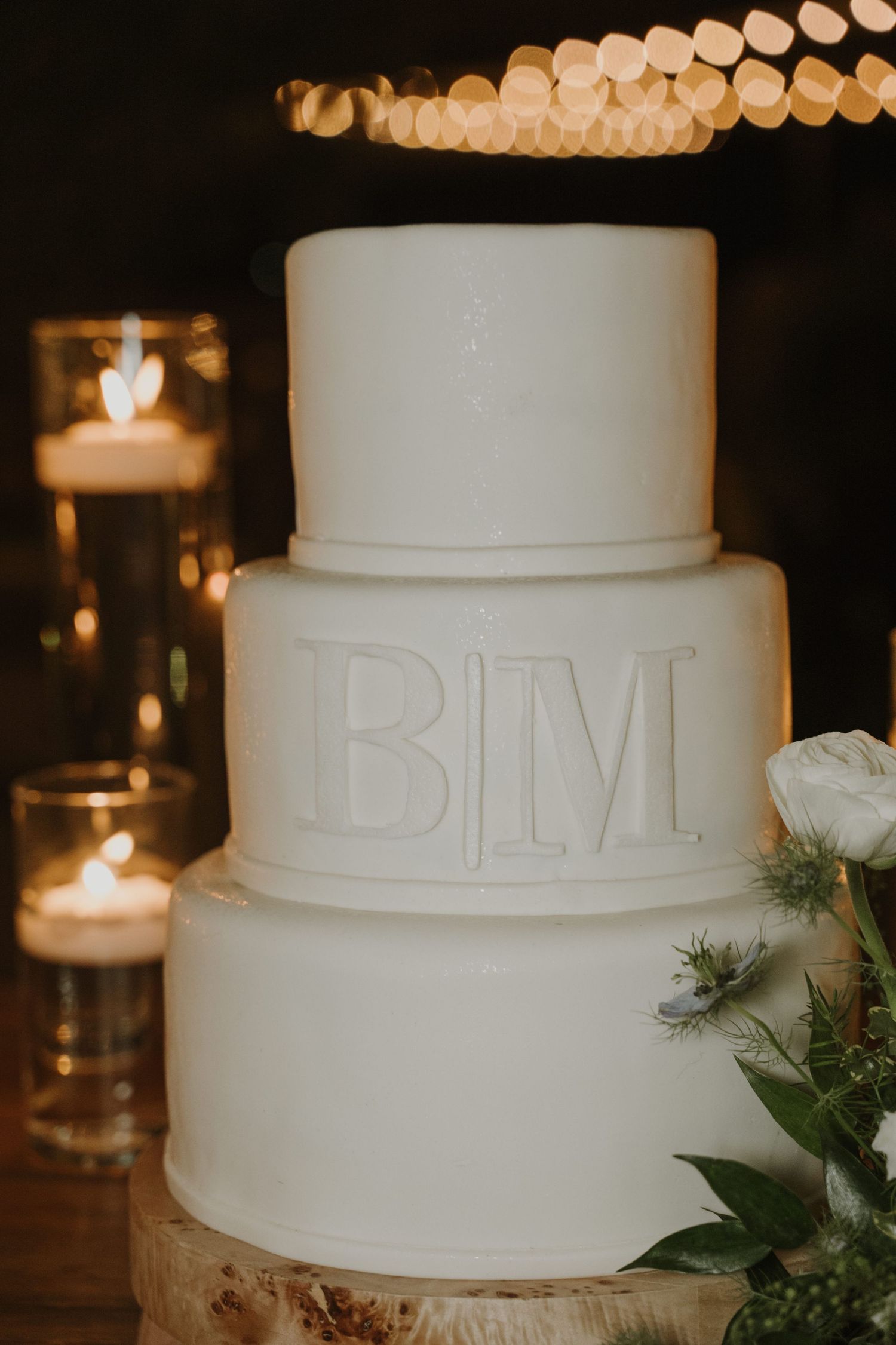 Elegant three-tier white wedding cake with monogram B|M initials, surrounded by glowing candles on a rustic wood slice.