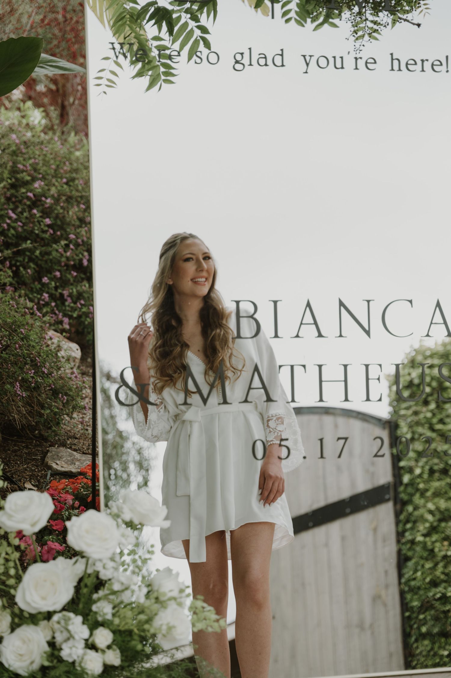 Bride in white robe smiling in front of wedding welcome sign reading Bianca and Matheus dated 05.17.