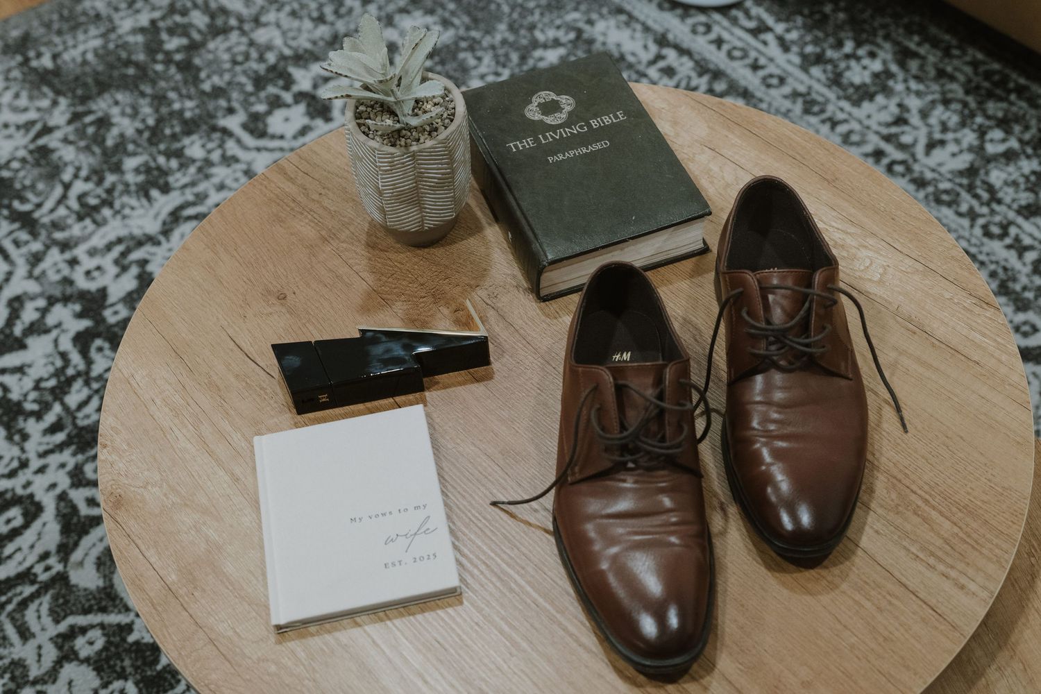 Brown leather dress shoes beside a Living Bible, cologne, and a wedding note card on a round wooden table.