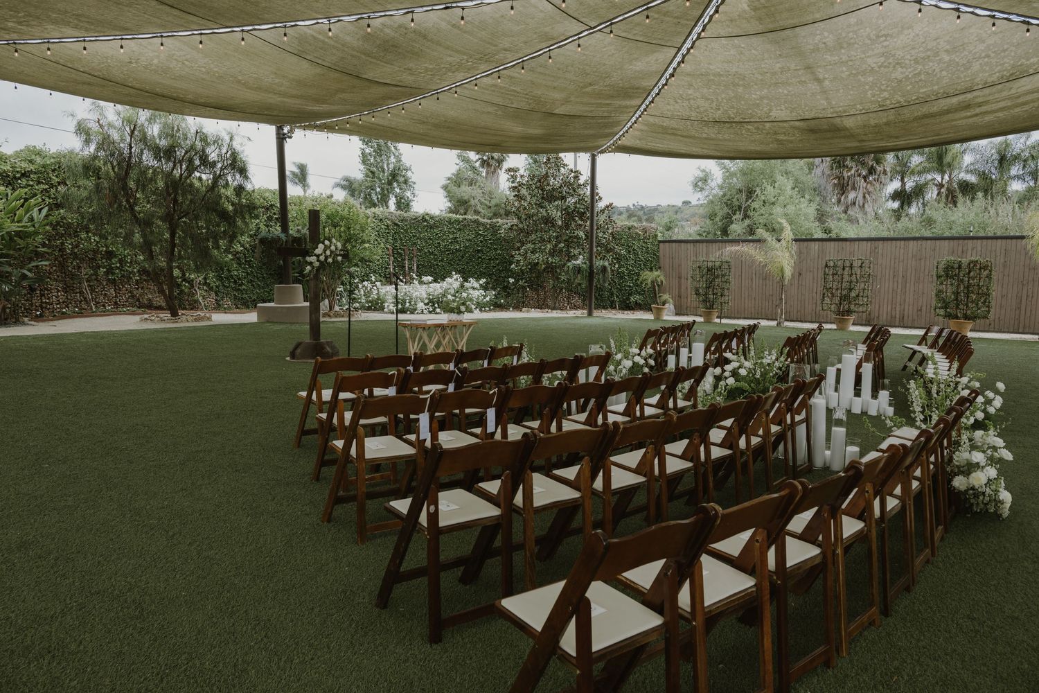 Elegant outdoor wedding ceremony setup with wooden folding chairs, floral arrangements, and a shaded tent on green lawn.