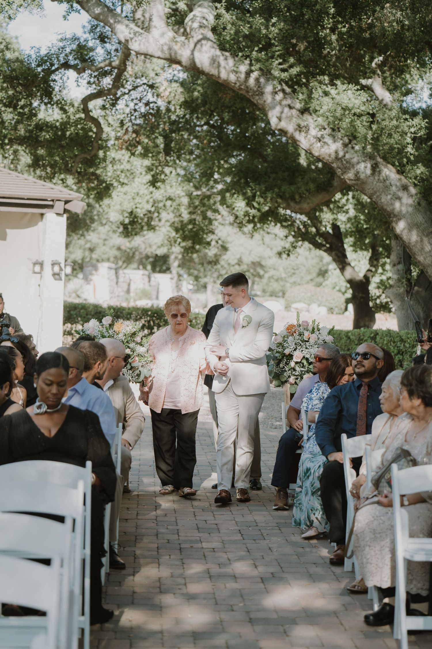 Groom in tan suit walks grandmother down outdoor wedding ceremony aisle past seated guests.
