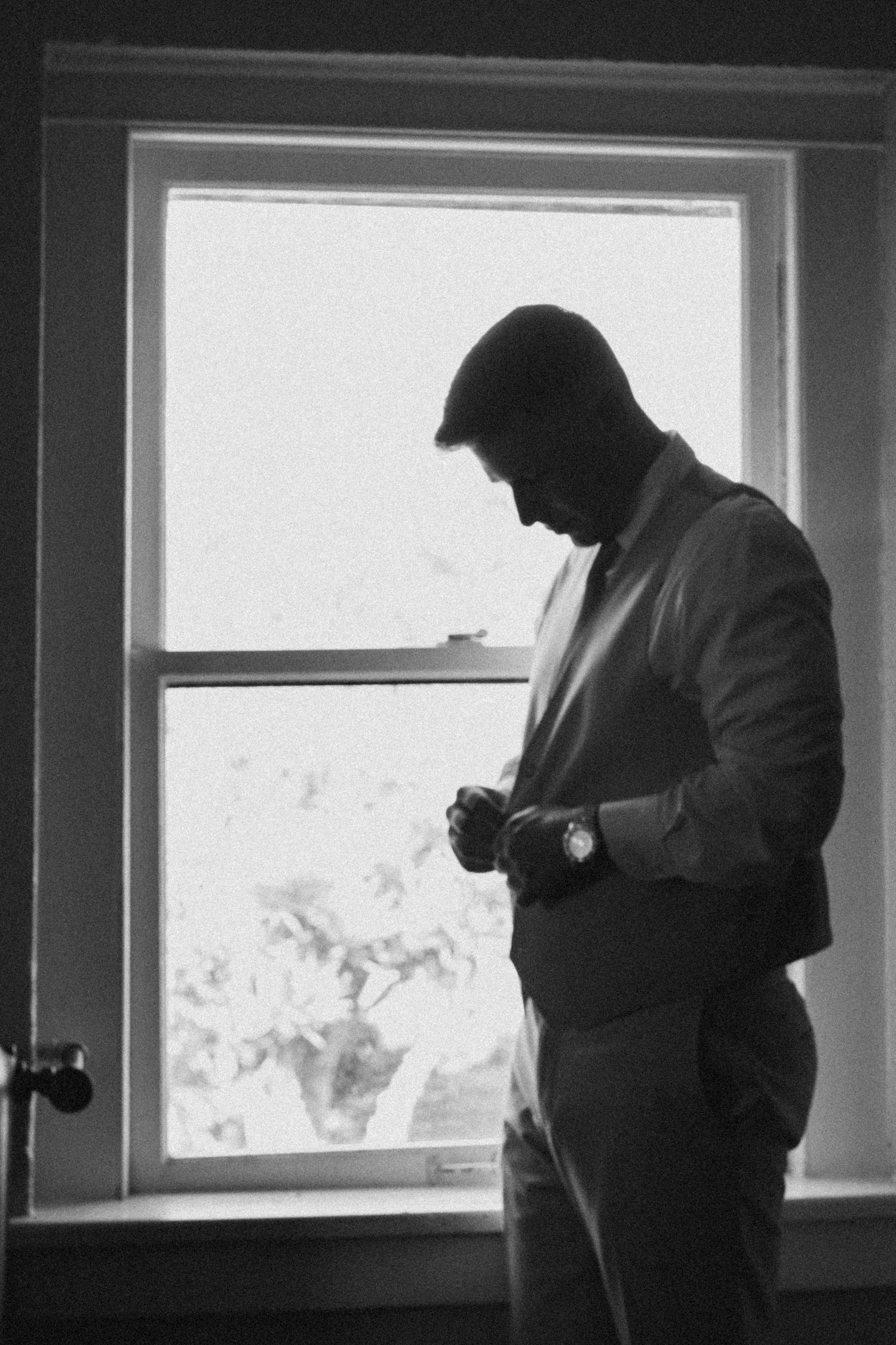 Black and white silhouette of a groom in a suit adjusting his cufflinks by a bright window.