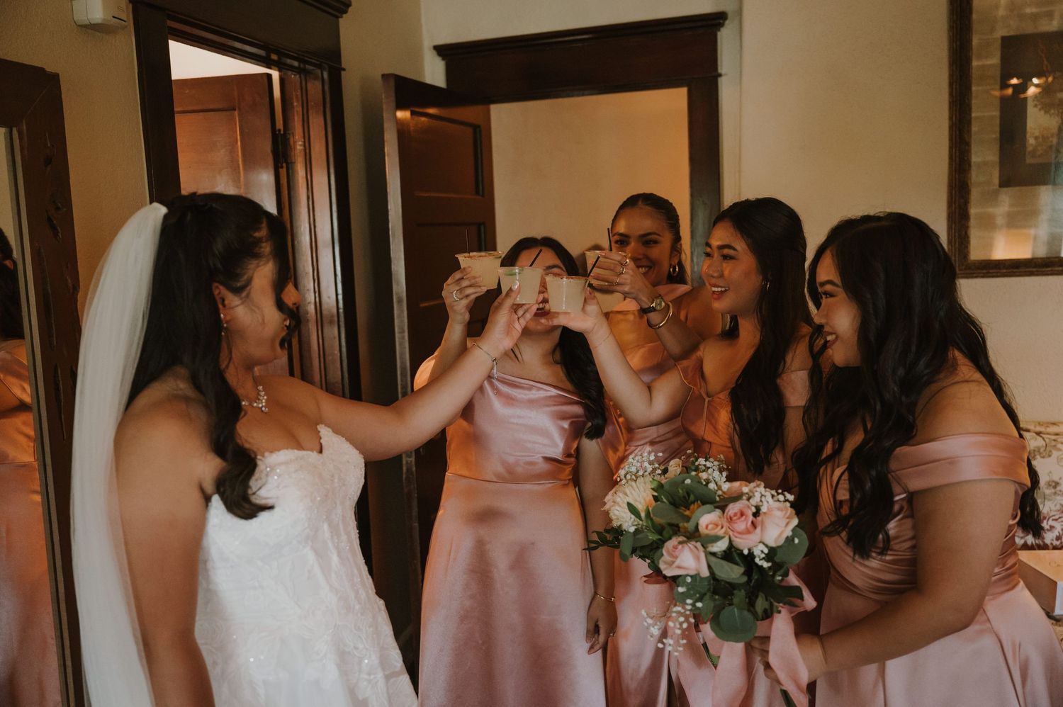 Bride and bridesmaids in pink satin dresses joyfully toasting together before the wedding.