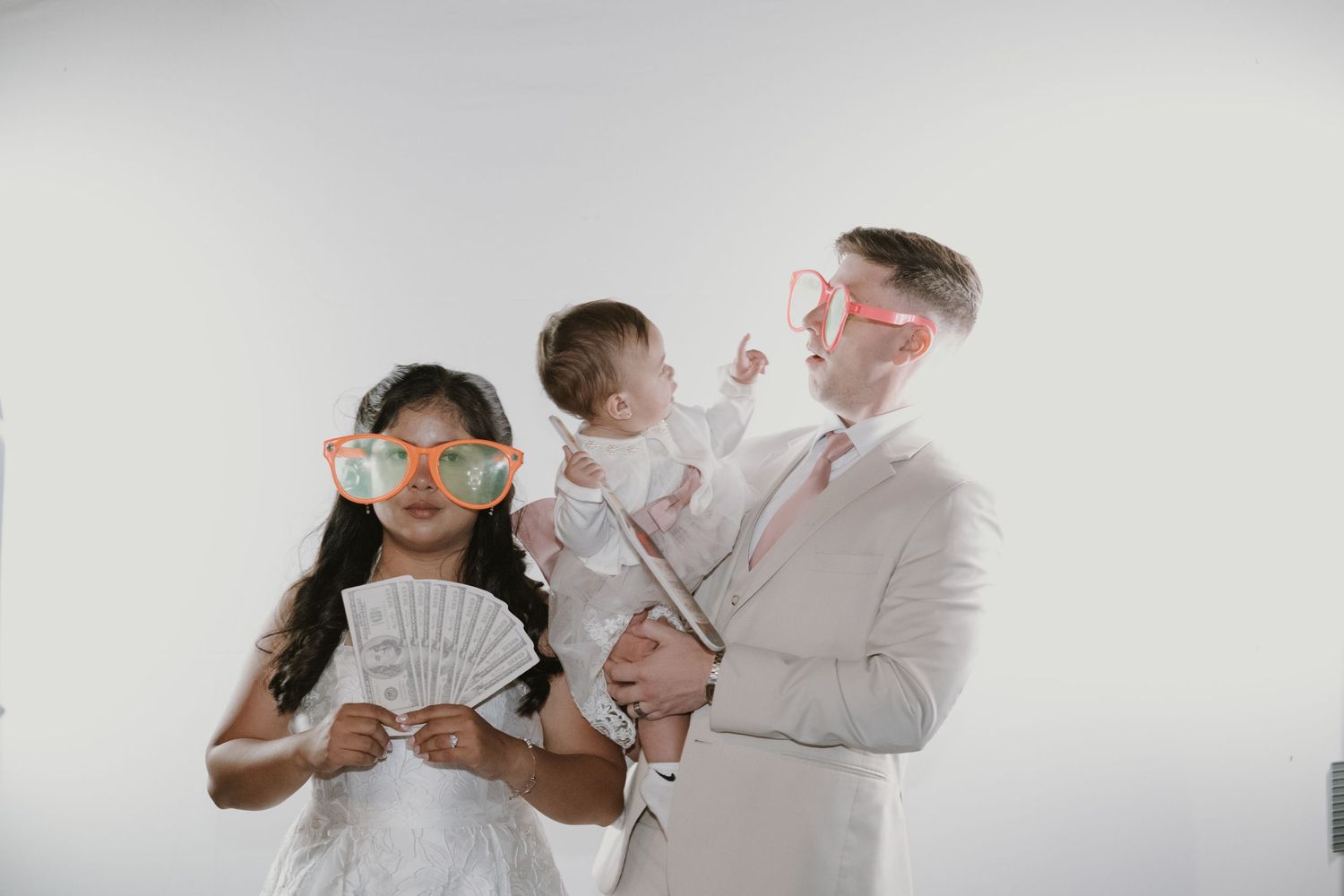 Family poses with fun oversized sunglasses and cash fan at wedding photo booth, man holding baby.