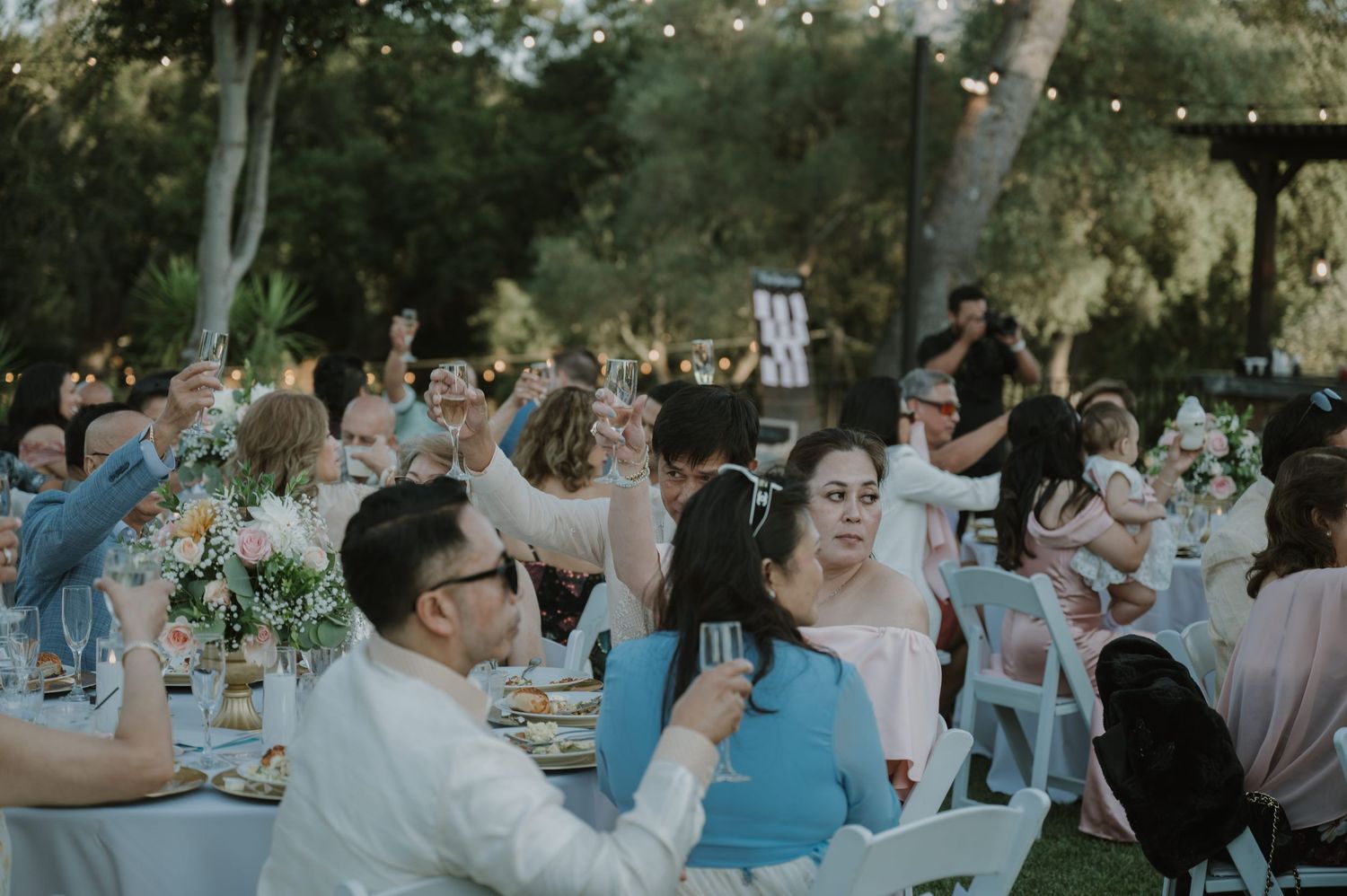 Guests raising glasses in a toast at an outdoor wedding reception surrounded by lush greenery and string lights.
