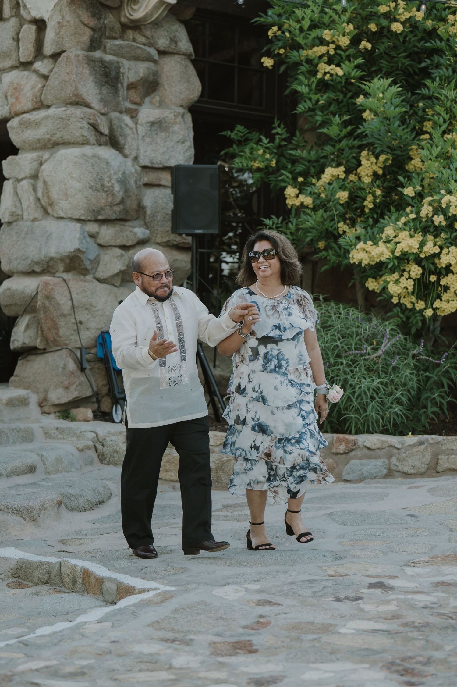 A man in a barong and woman in a floral dress walk together at an outdoor wedding reception.