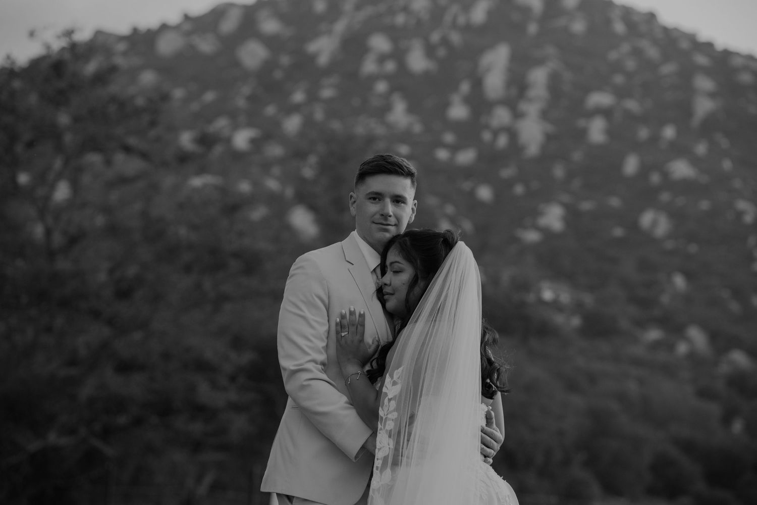 Groom embraces bride in a tender black and white wedding portrait with mountain backdrop.