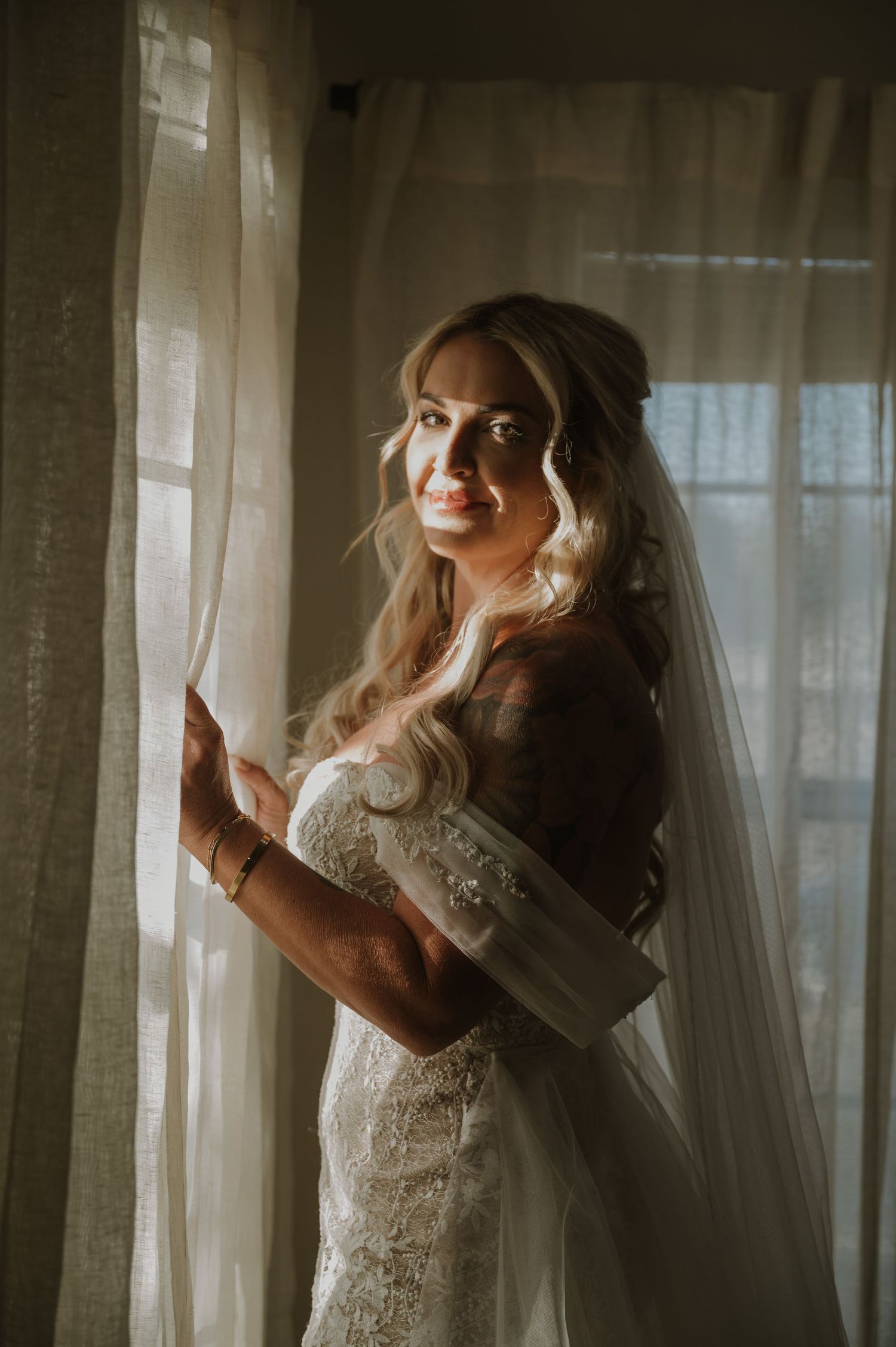 A bride in a lace wedding dress poses elegantly by sheer curtains in soft natural lighting.