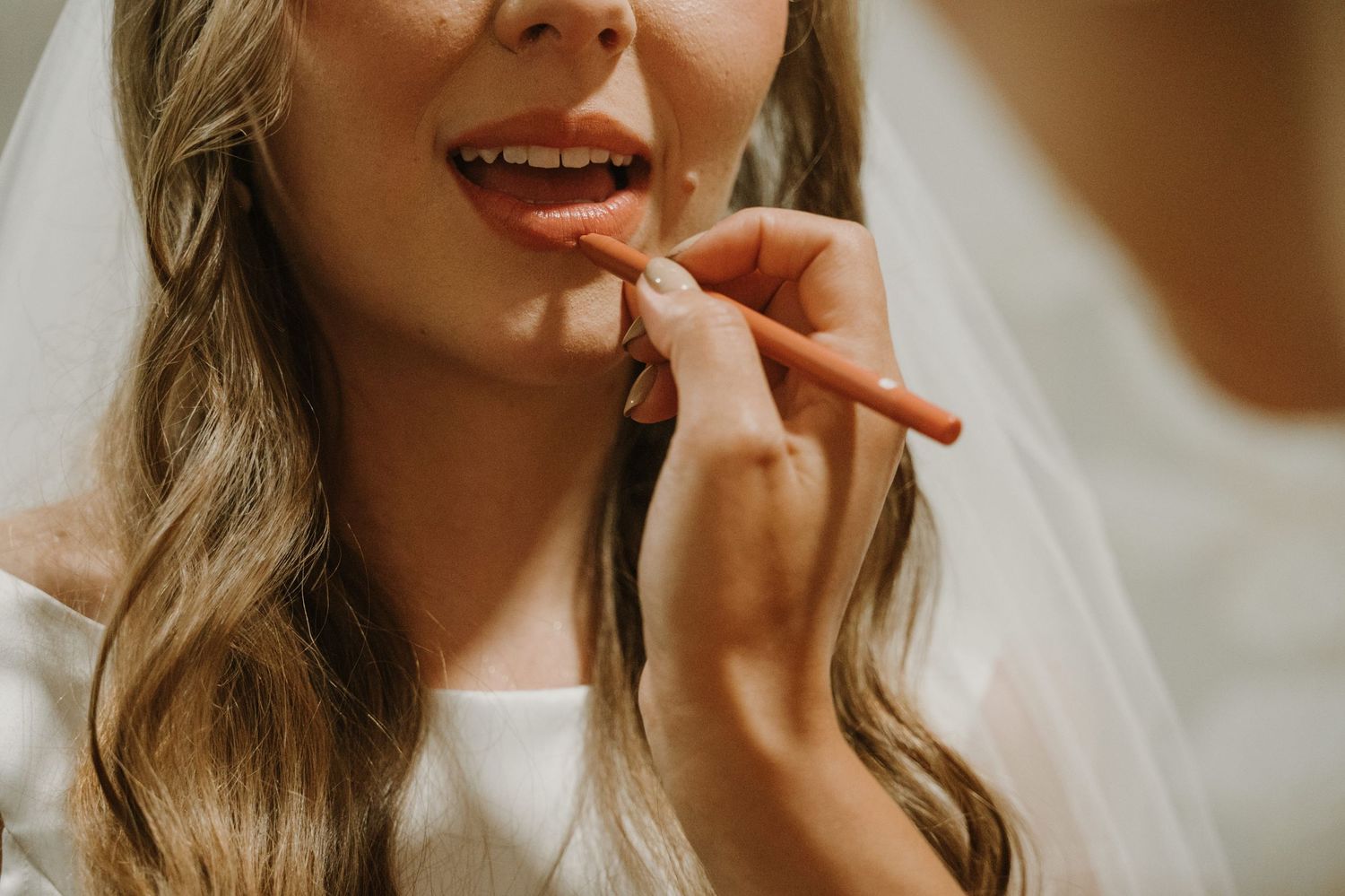 Bride applying coral lipstick while wearing a white veil, preparing for her wedding day.