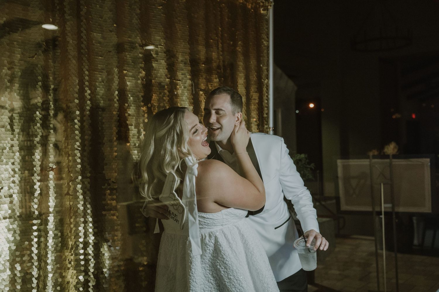 Bride and groom sharing a romantic embrace in front of a glittering gold sequin backdrop at their wedding reception.