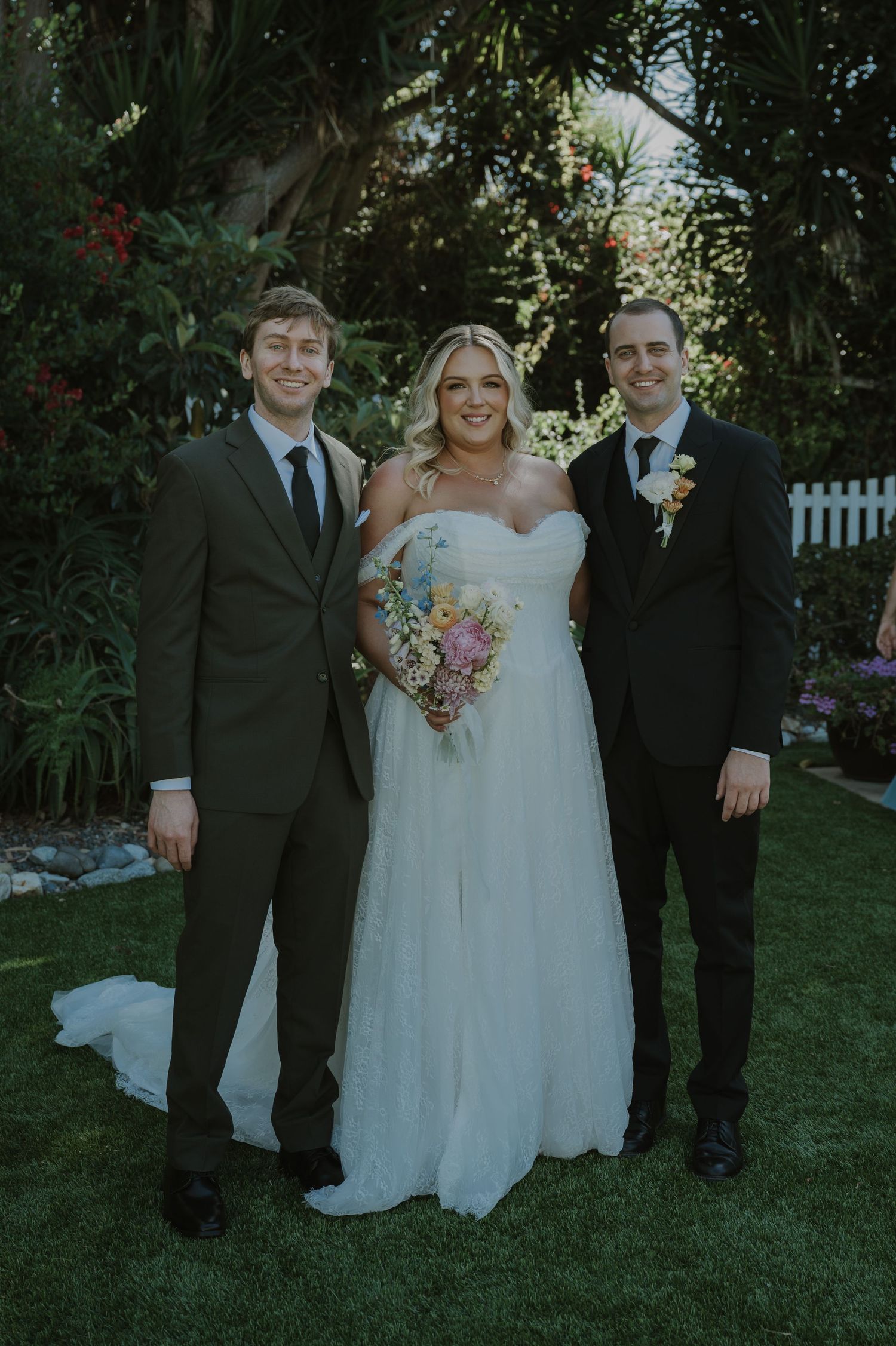 Bride in white gown holds colorful bouquet flanked by two groomsmen in suits at outdoor garden wedding.