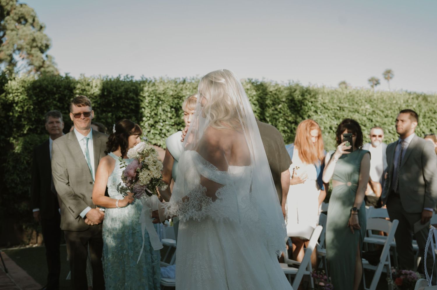 Bride walks down outdoor wedding aisle holding bouquet, guests seated in white chairs, lush green hedges backdrop.