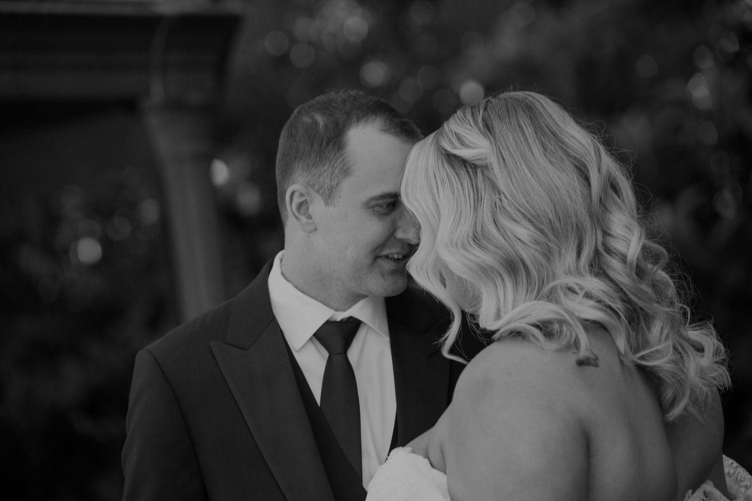 Black and white wedding photo of smiling groom in suit facing blonde bride outdoors.
