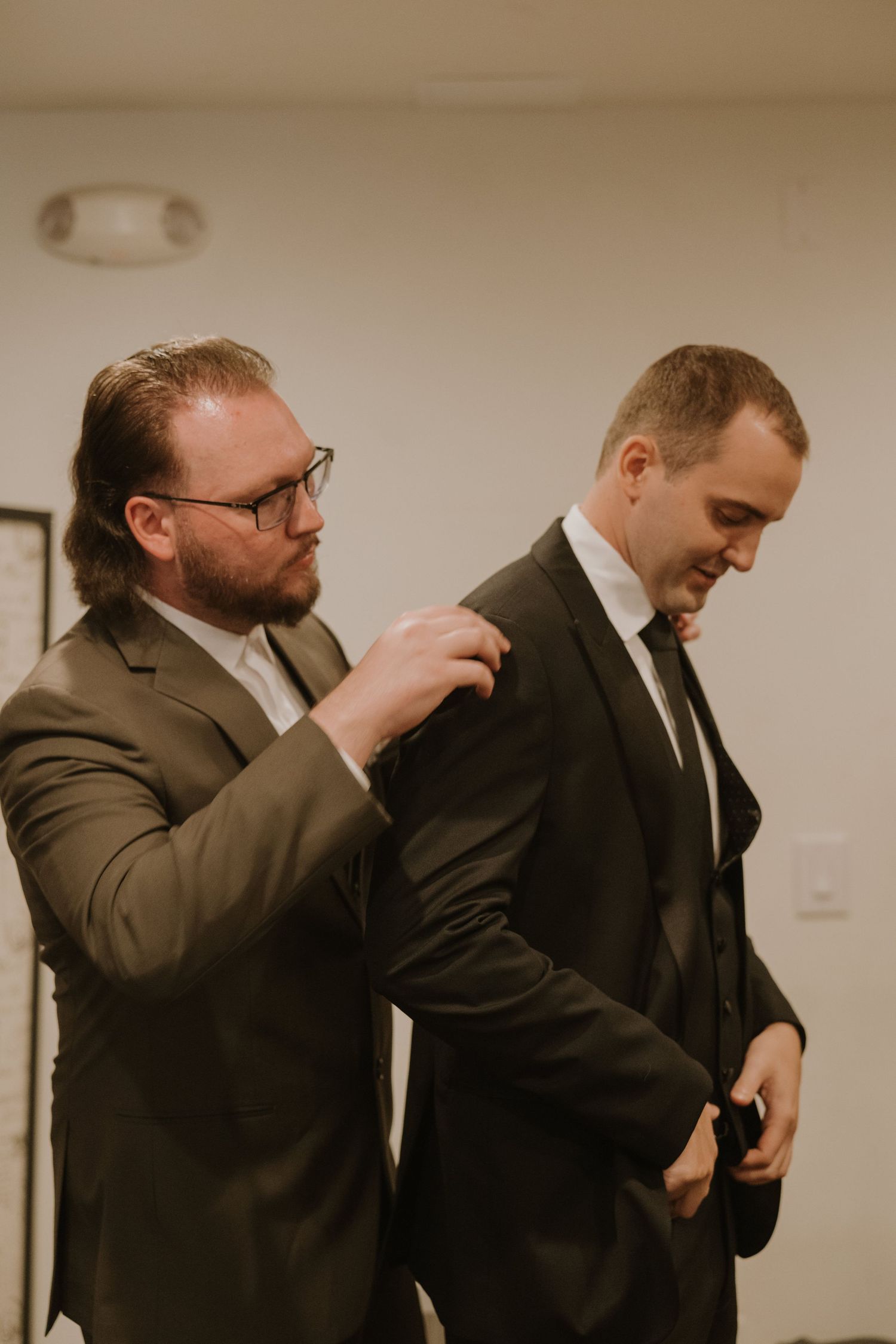 Two men in dark suits prepare for a wedding ceremony, one helping the other adjust his jacket.