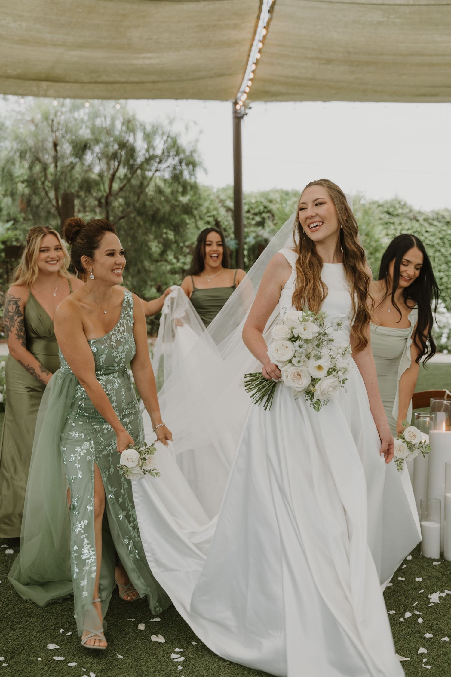 Bride in white gown laughs with bridesmaids in sage green dresses at outdoor wedding reception.