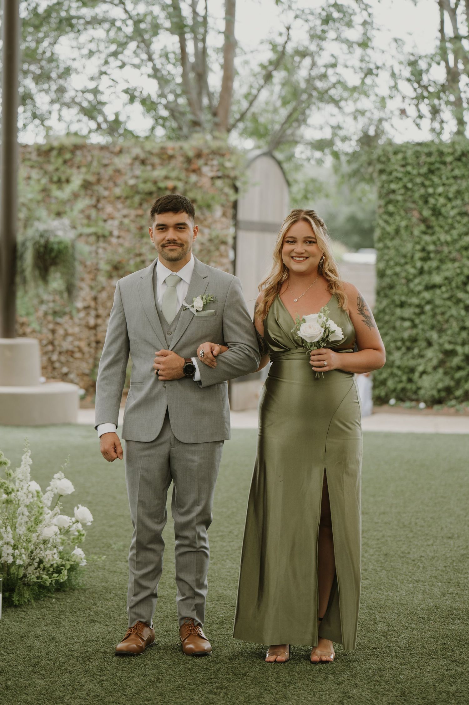 A groomsman in a gray suit and bridesmaid in an olive green gown pose together at an outdoor wedding ceremony.