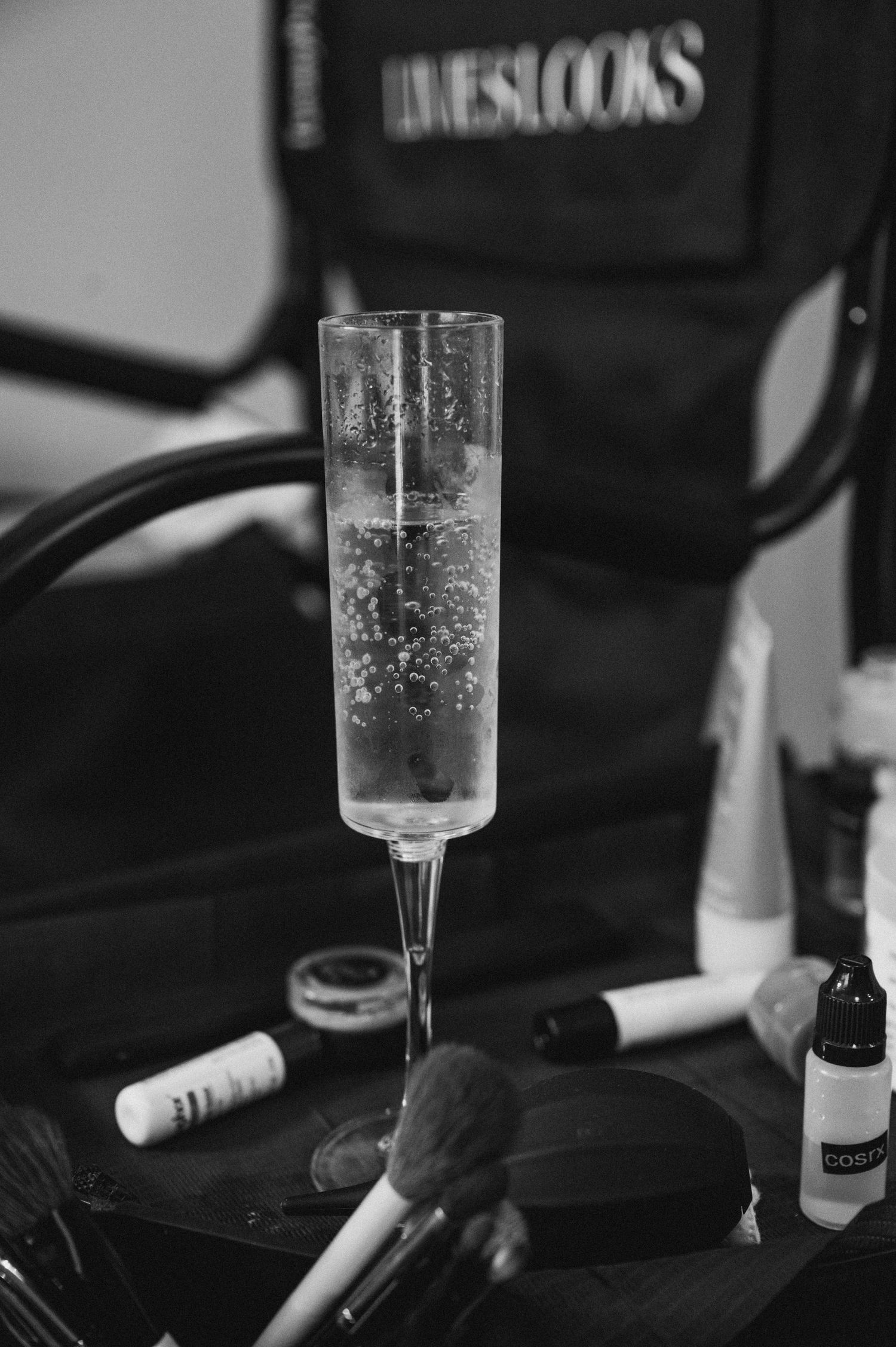 Black and white photo of champagne flute surrounded by makeup brushes and beauty products on a vanity table.