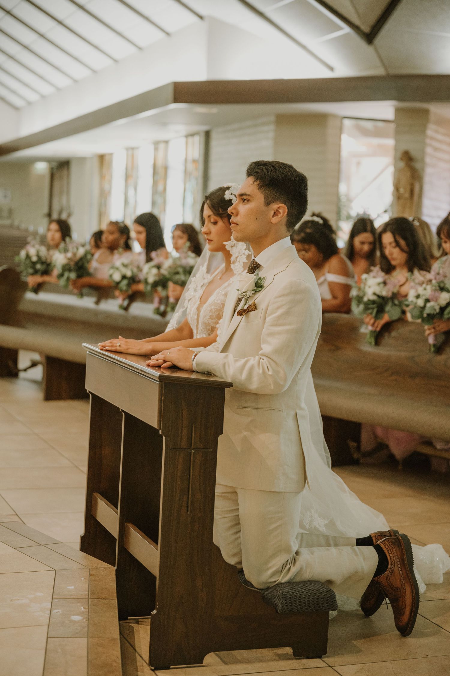 Groom in white suit kneels at church prie-dieu during wedding ceremony, bridesmaids seated in pews behind.
