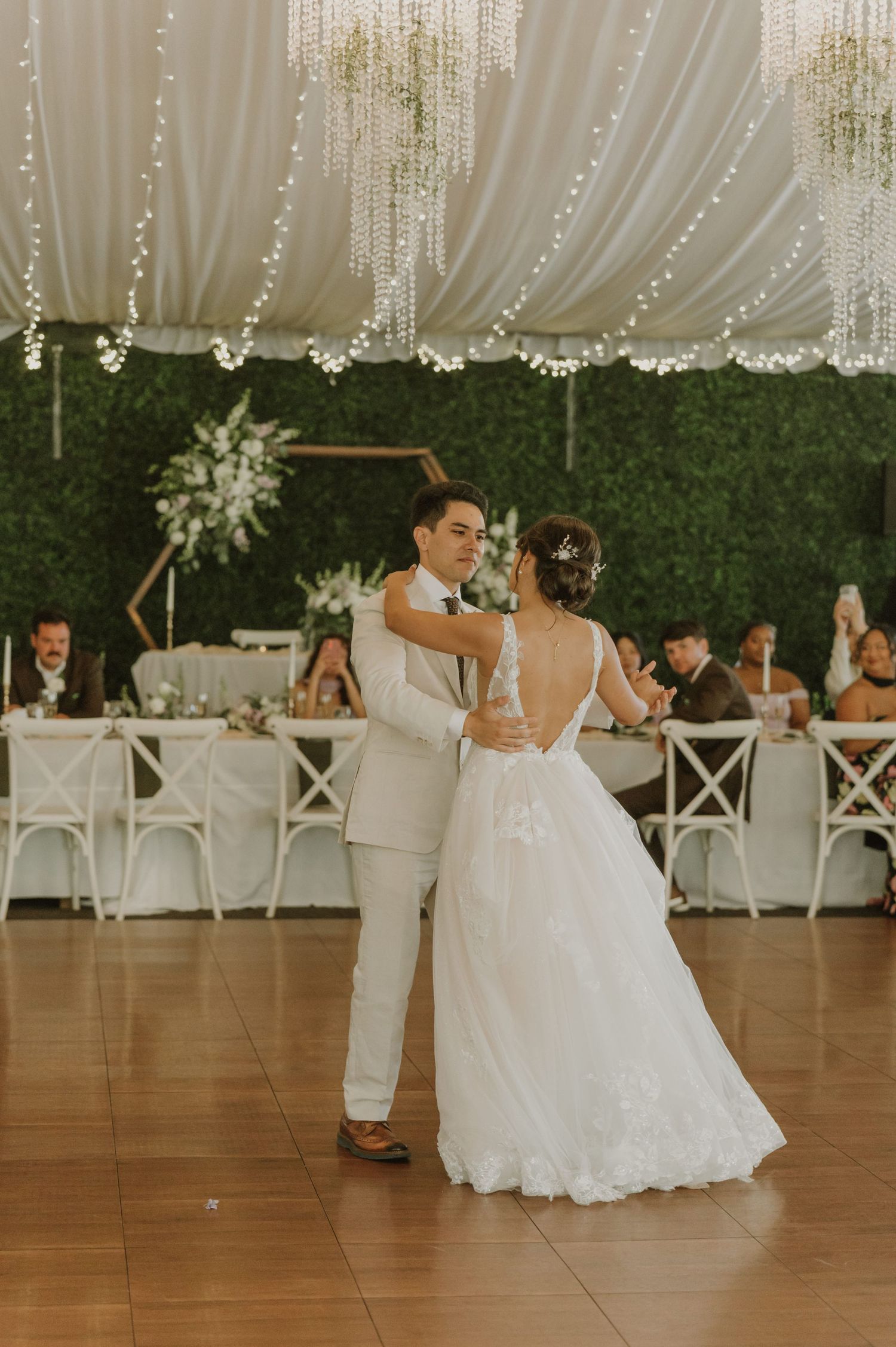 Bride and groom share their first dance in an elegant wedding venue with draped white fabric and twinkling lights.