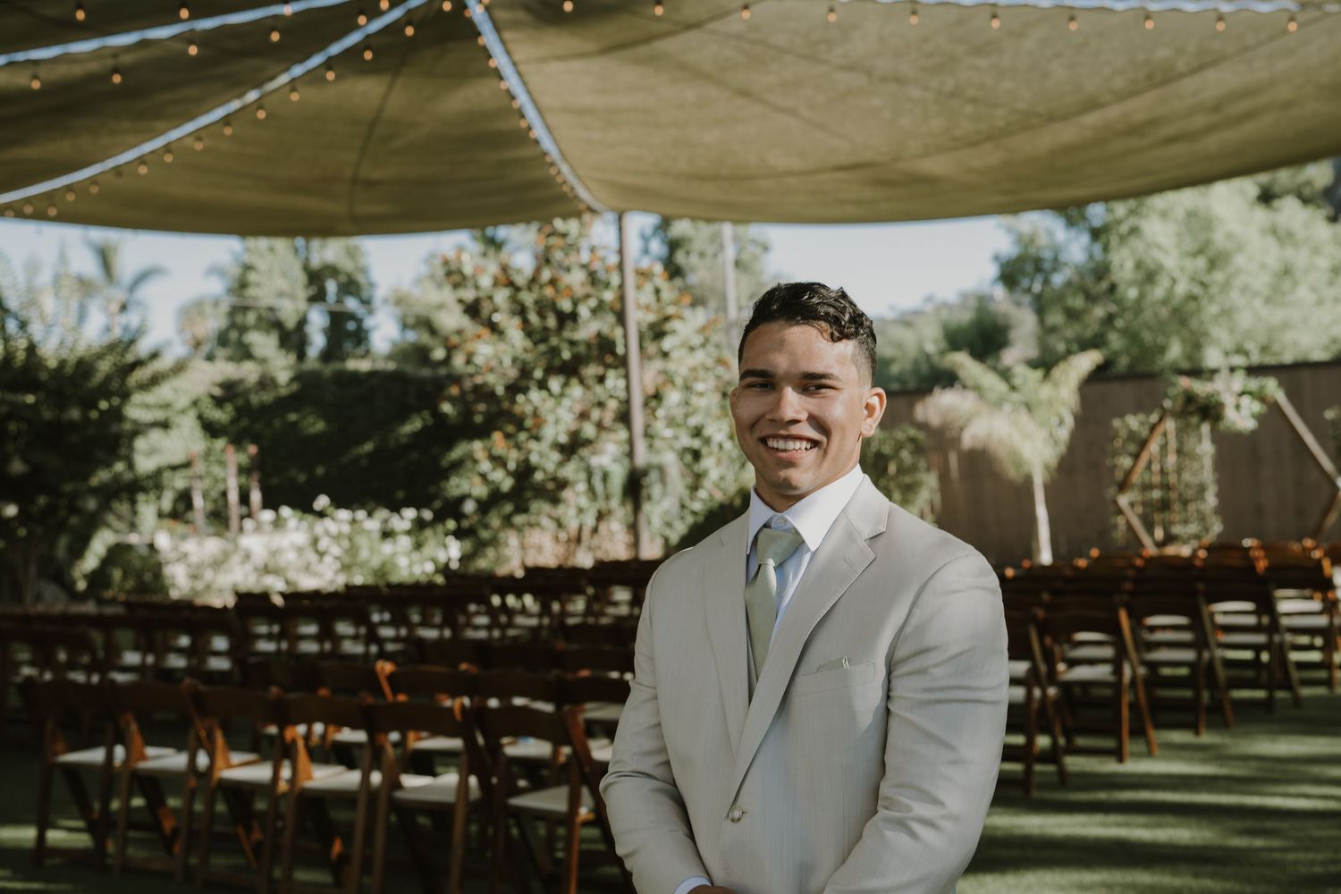 Smiling groom in a light beige suit standing at an outdoor wedding ceremony venue with rows of chairs.